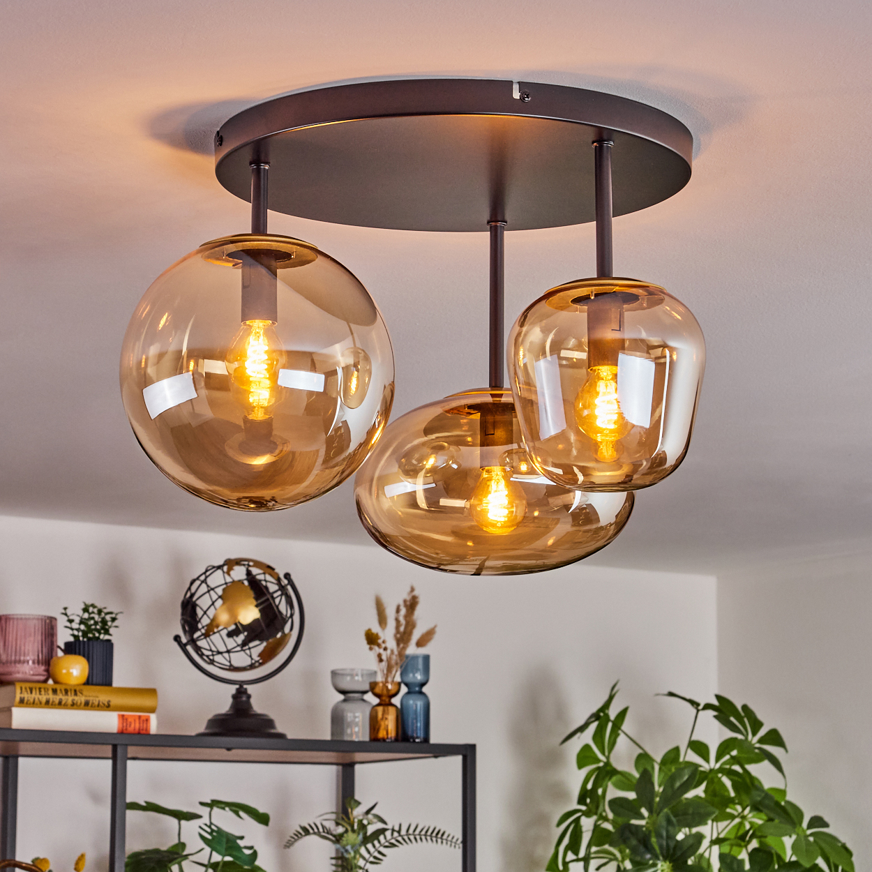 Eine Deckenlampe mit drei bernsteinfarbenen Glasschirmen. Darunter ein Regal mit Dekoration, darunter ein Globus und Pflanzen.