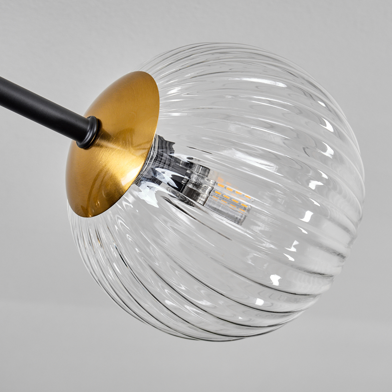 Nahaufnahme einer klaren, gerippten Glaskugel-Leuchte mit goldener Kappe und schwarzem Arm vor weißem Hintergrund.