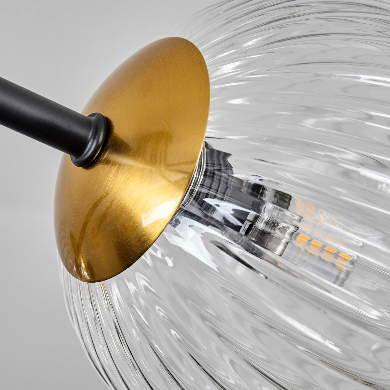 Nahaufnahme einer Glaslampe. Die Lampe hat einen goldenen Sockel, einen schwarzen Stiel und einen durchsichtigen, geriffelten Glasschirm. Der Hintergrund ist schlicht weiß.