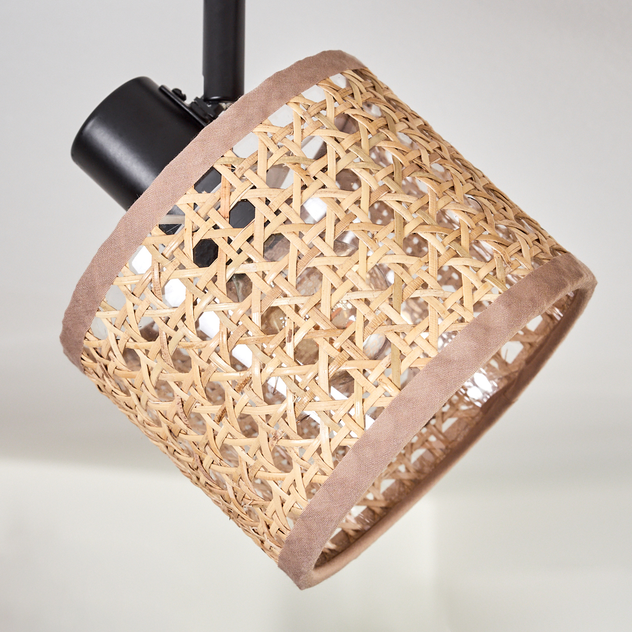 Ein Lampenschirm aus gewebtem Rattan mit brauner Zierleiste, der an einer schwarzen Metallhalterung vor weißem Hintergrund hängt.