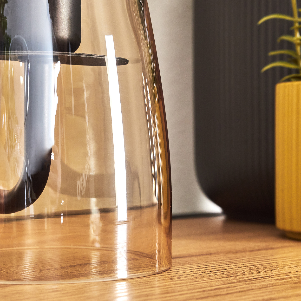 Ein klares, bernsteinfarbenes Glas steht auf einem Holztisch neben einer dunklen Vase und einem gelben Topf mit einer Pflanze.