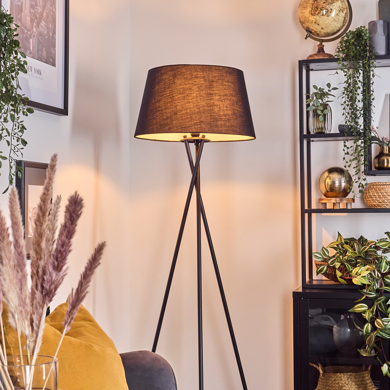 Eine schwarze Stehlampe mit Schirm steht in einem Raum. Ein Regal mit Pflanzen und Dekoration ist sichtbar. Eine Vase mit Pampasgras ist im Vordergrund.