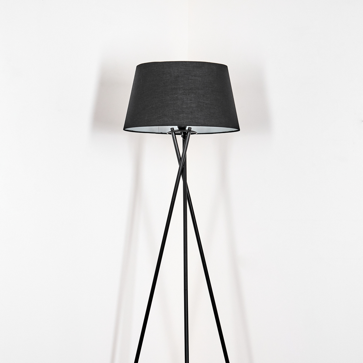 Eine schwarze Stehlampe steht in einer Ecke vor einer weißen Wand. Die Lampe hat einen schwarzen Schirm und ein Dreibein.