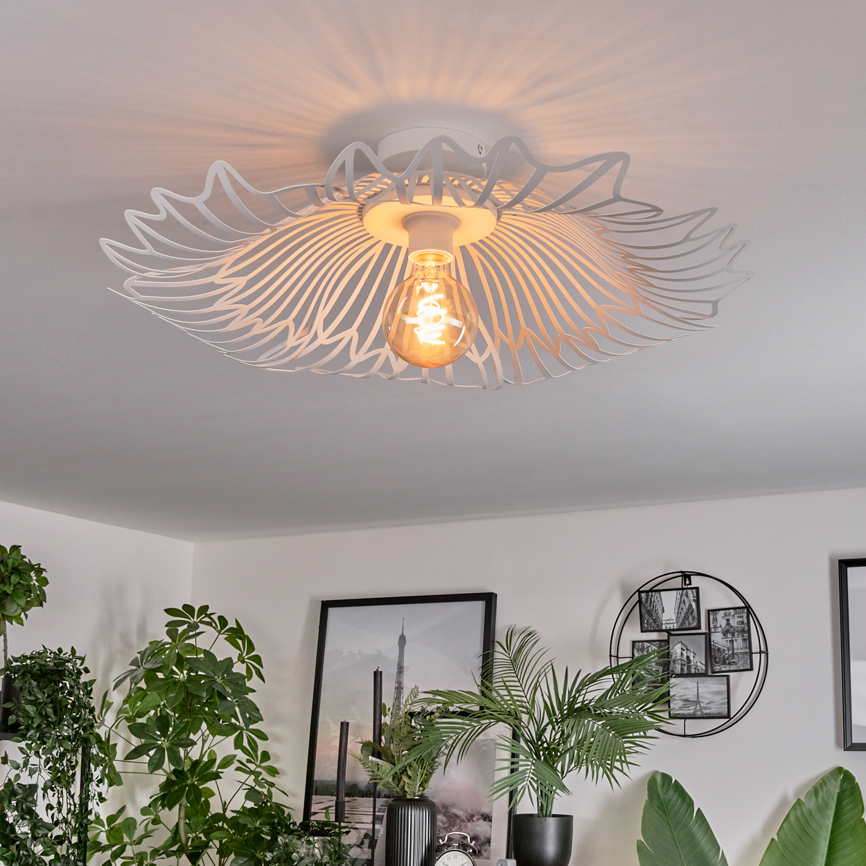 Eine weiße Deckenlampe mit floralem Design und leuchtender Glühbirne. Darunter Pflanzen, gerahmte Bilder und eine Uhr.