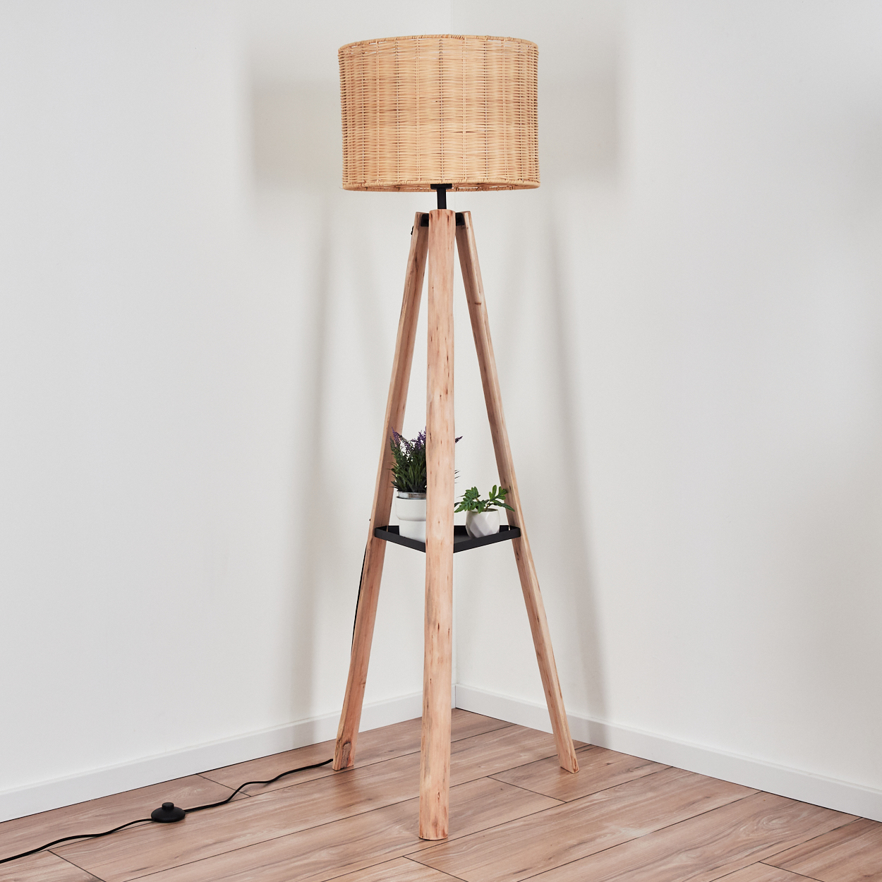 Eine hölzerne Stativ-Stehlampe mit gewebtem Schirm. Sie hat ein Regal mit zwei Topfpflanzen. Die Lampe steht auf einem Holzboden.