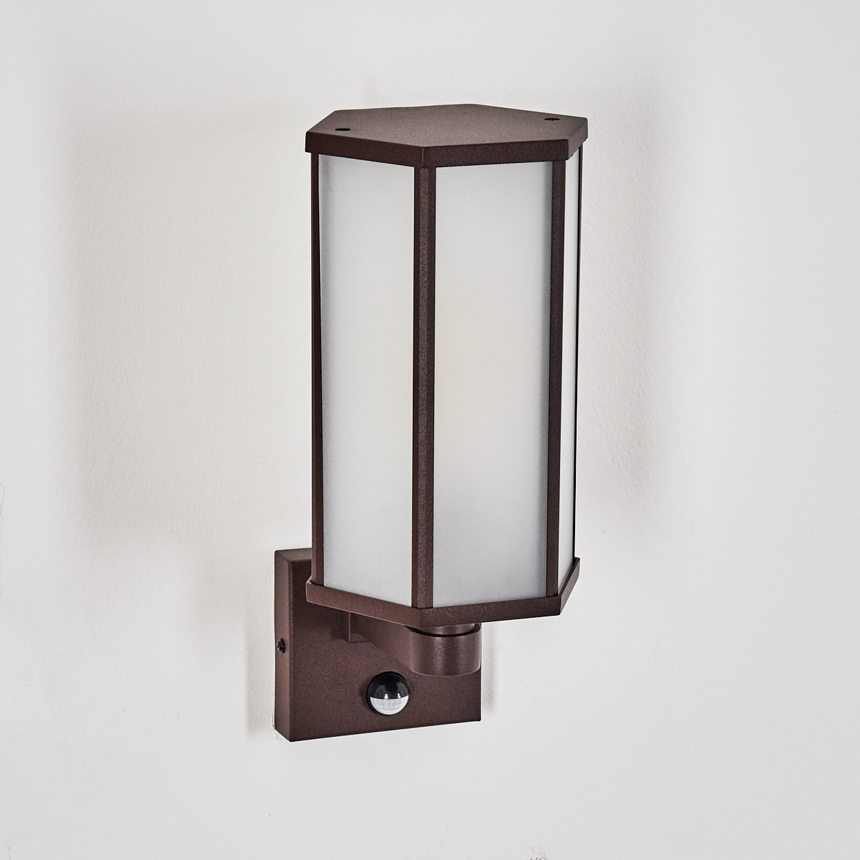 Eine braune Außenwandleuchte mit weißem, mattiertem Glas. Sie hat einen Bewegungssensor und ist an einer weißen Wand montiert.