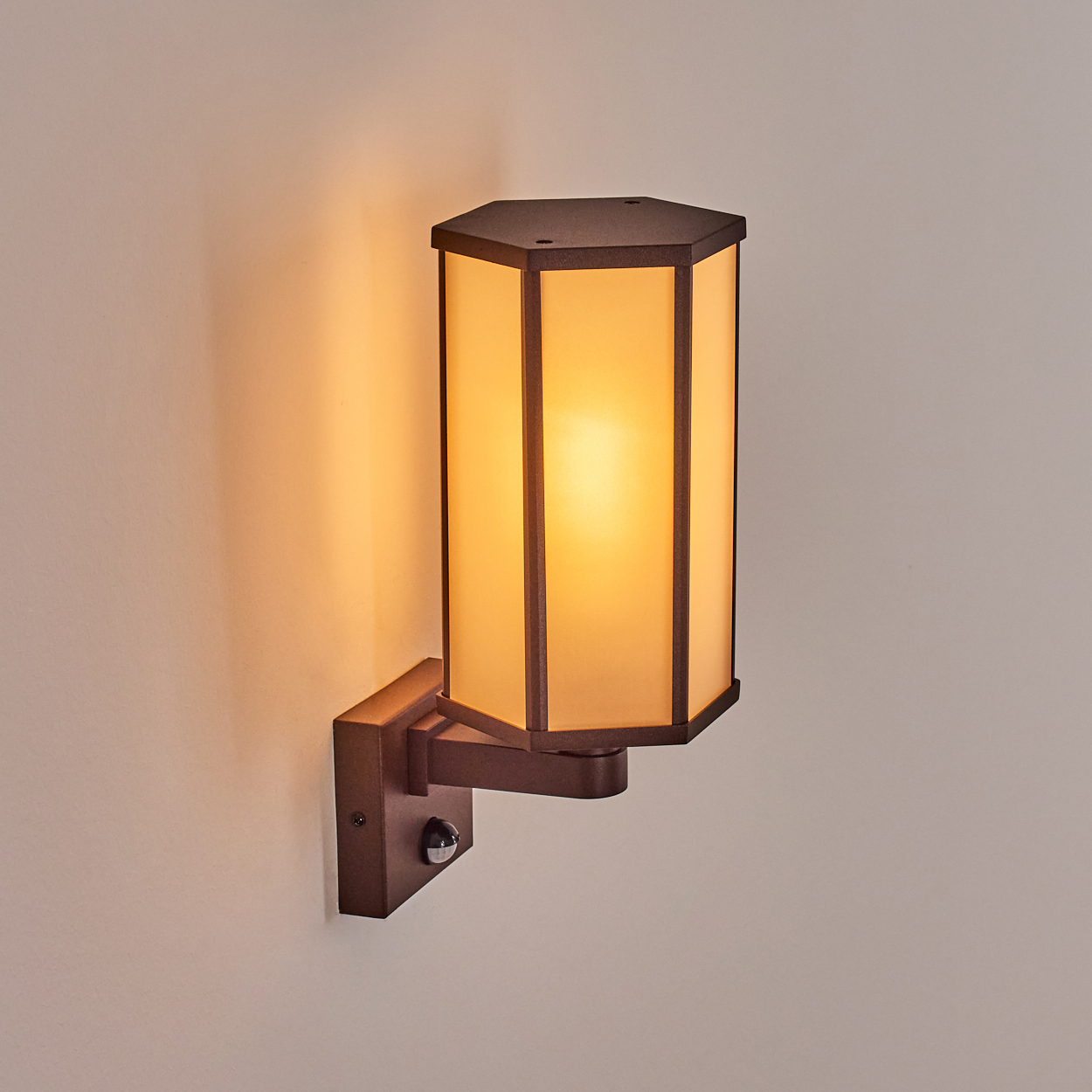 Eine wandmontierte, bronzefarbene Lampe mit einem beleuchteten, rechteckigen Schirm. Die Lampe ist an einer hellen Wand befestigt.