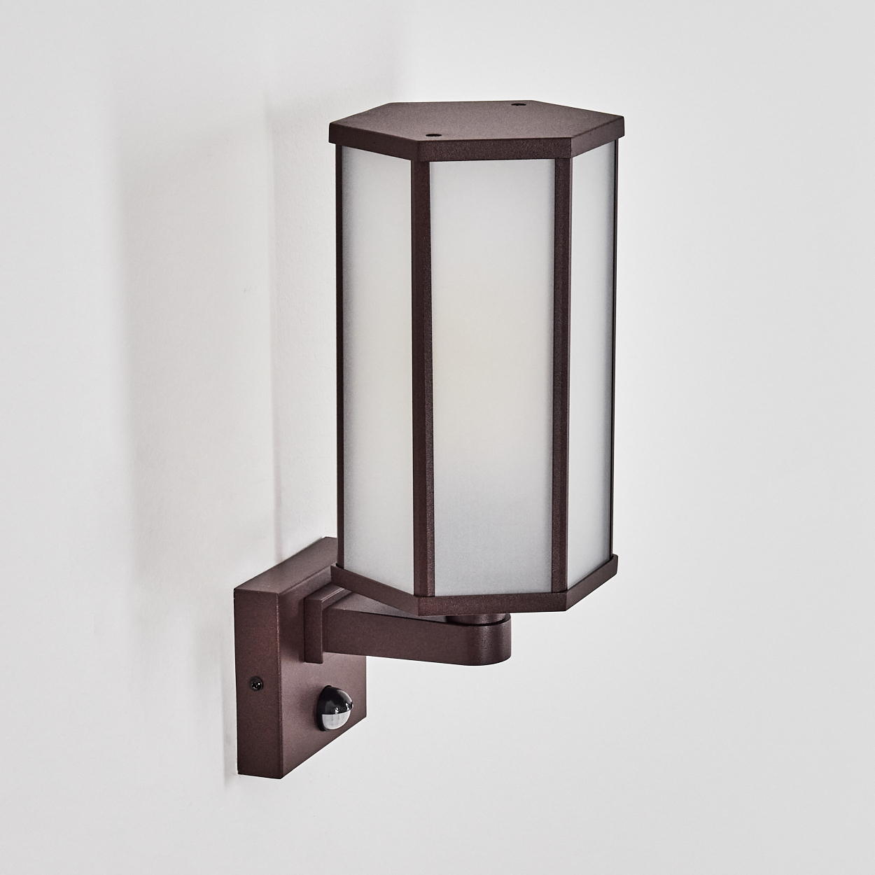 Eine braune, sechseckige Außenwandleuchte mit einem Schirm aus Milchglas. Sie hat einen Bewegungssensor und ist an einer weißen Wand befestigt.