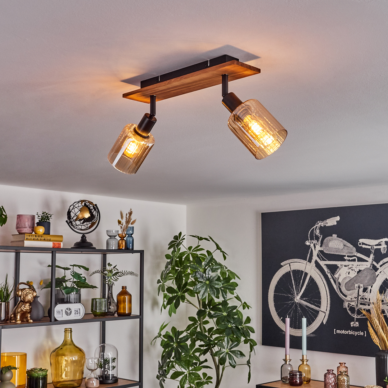 Deckenleuchte mit zwei Lampen, Holzsockel. Dekoartikel auf Regalen, eine Pflanze und ein Motorradkunstwerk sind sichtbar.