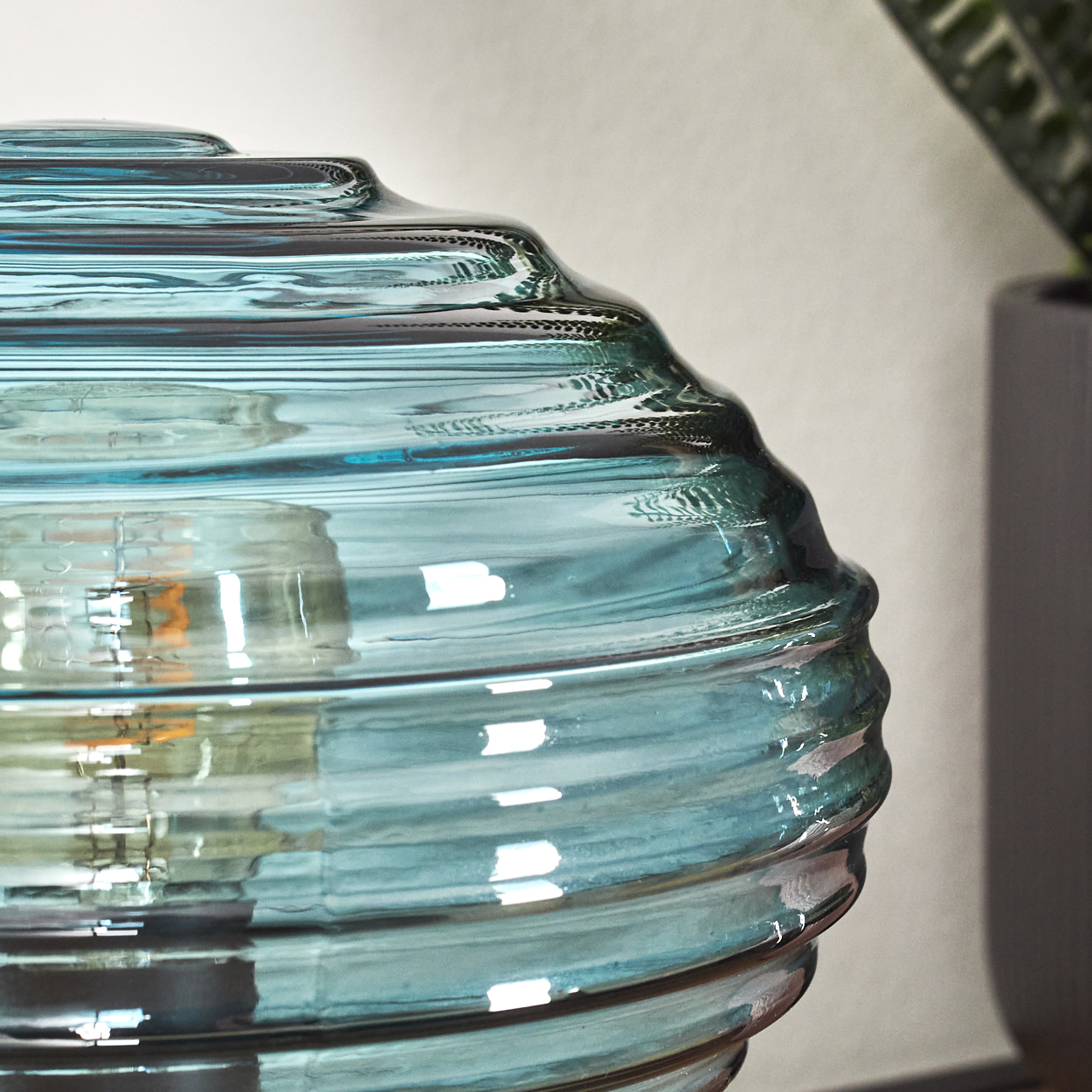 Nahaufnahme einer tealfarbenen Glasvase mit geriffelter Oberfläche. Im Hintergrund eine Pflanze. Die Vase reflektiert das Licht.