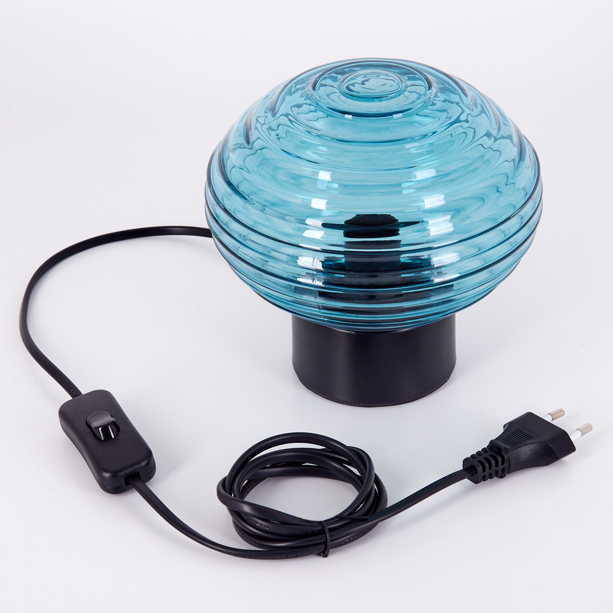Eine blaue Glaslampe mit schwarzem Sockel und Kabel. Die Lampe steht auf einer weißen Oberfläche. Ein schwarzer Netzstecker ist ebenfalls sichtbar.