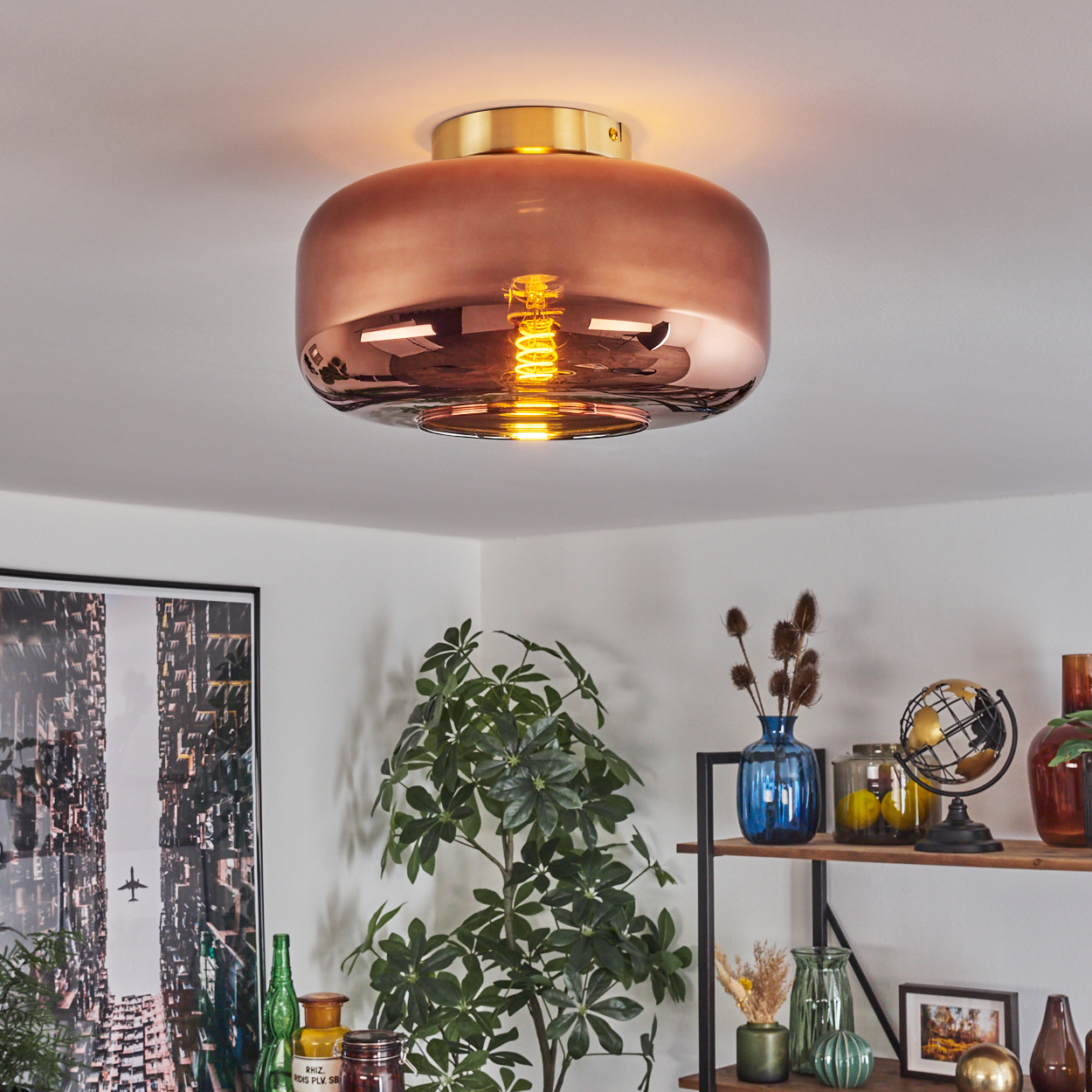Kupferfarbene Deckenlampe mit freiliegender Glühbirne. Darunter ein Regal mit Deko, Pflanzen und einem Bild an der Wand.