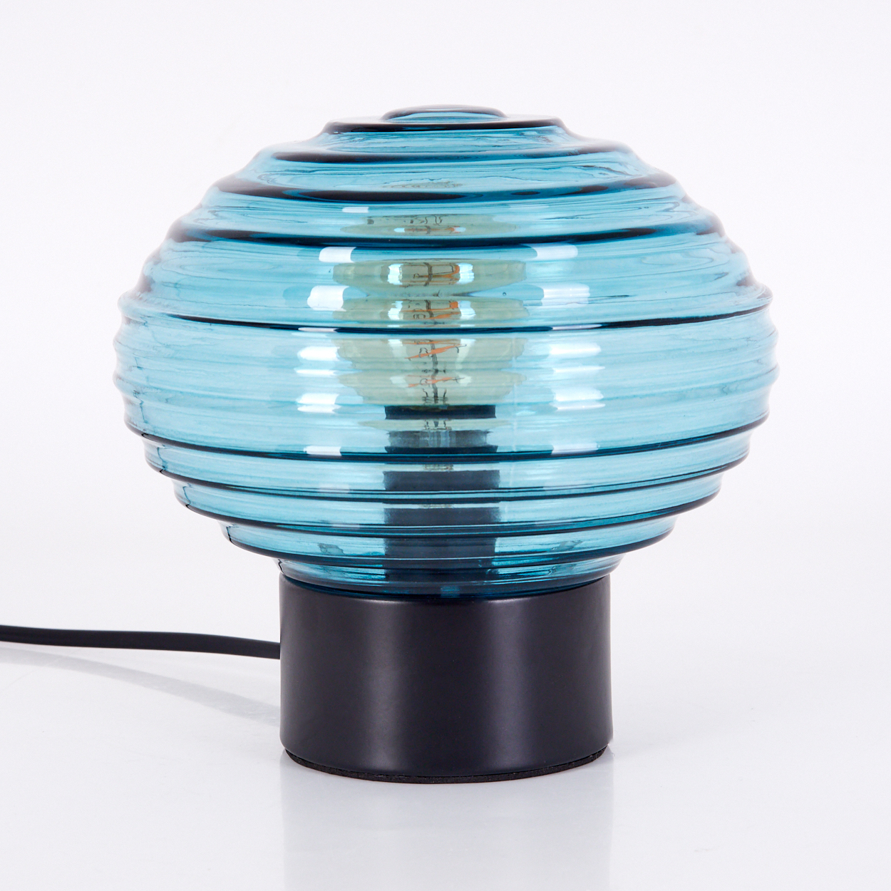 Eine blaue Glaslampe mit schwarzem Sockel und einer sichtbaren Glühbirne. Die Lampe steht auf einer weißen Oberfläche.
