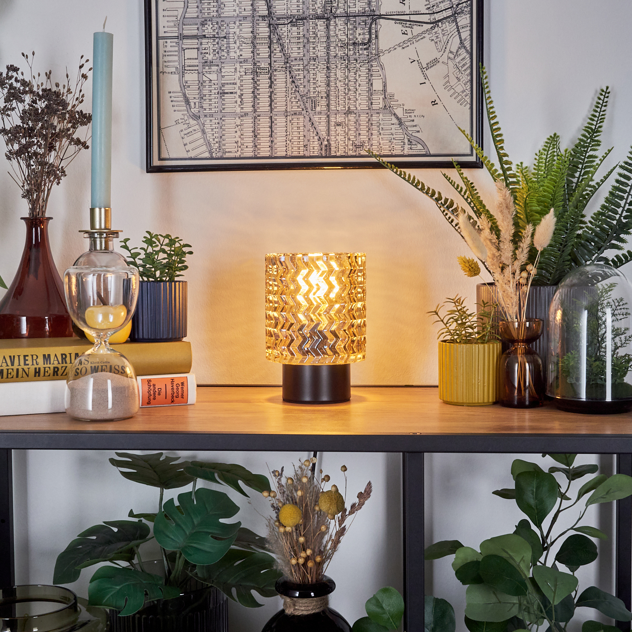 Ein Tisch mit Lampe, Pflanzen, Büchern und einer Karte. Die Lampe leuchtet und hat einen gemusterten Schirm. Es gibt auch eine Kerze.