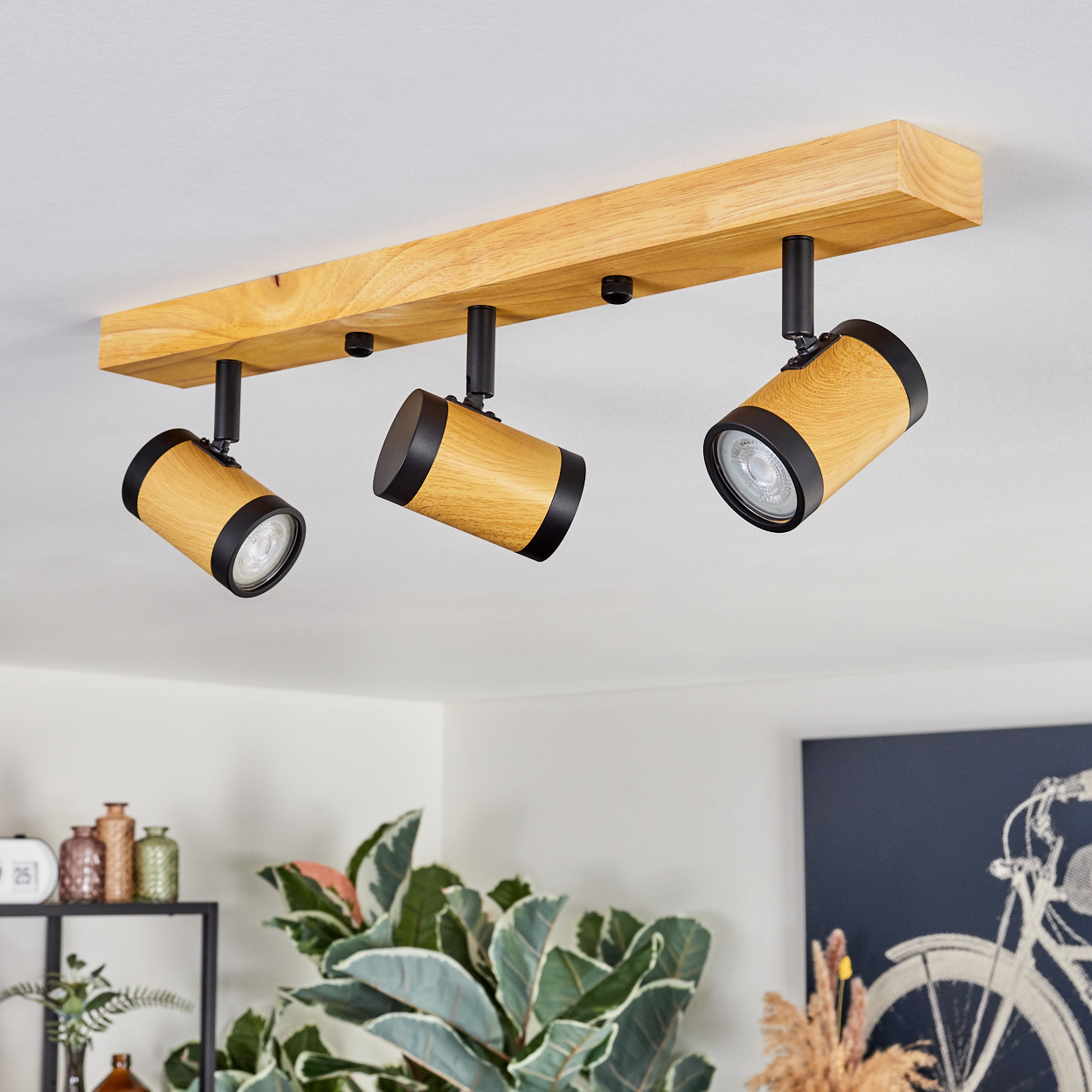 Drei Deckenleuchten aus Holz mit schwarzen Akzenten. Die Lampen sind an einem Holzbalken befestigt. Hintergrund zeigt Pflanzen und ein Gemälde.