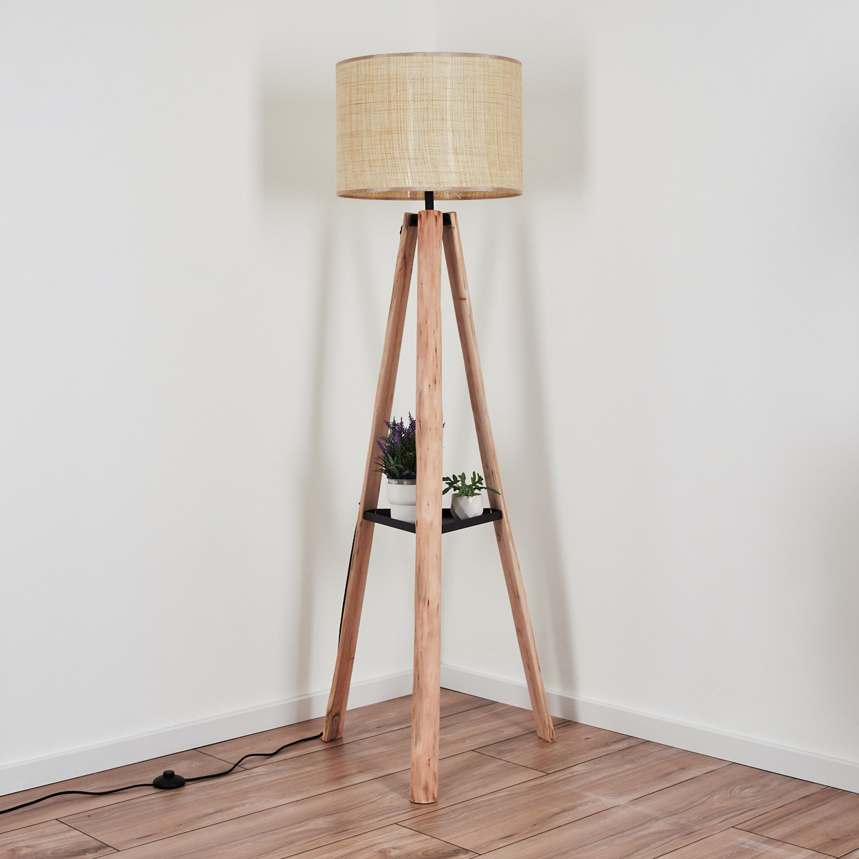 Eine hölzerne Stativ-Stehlampe mit beigem Lampenschirm. Sie hat ein Regal mit Pflanzen. Der Raum hat einen Holzboden und weiße Wände.
