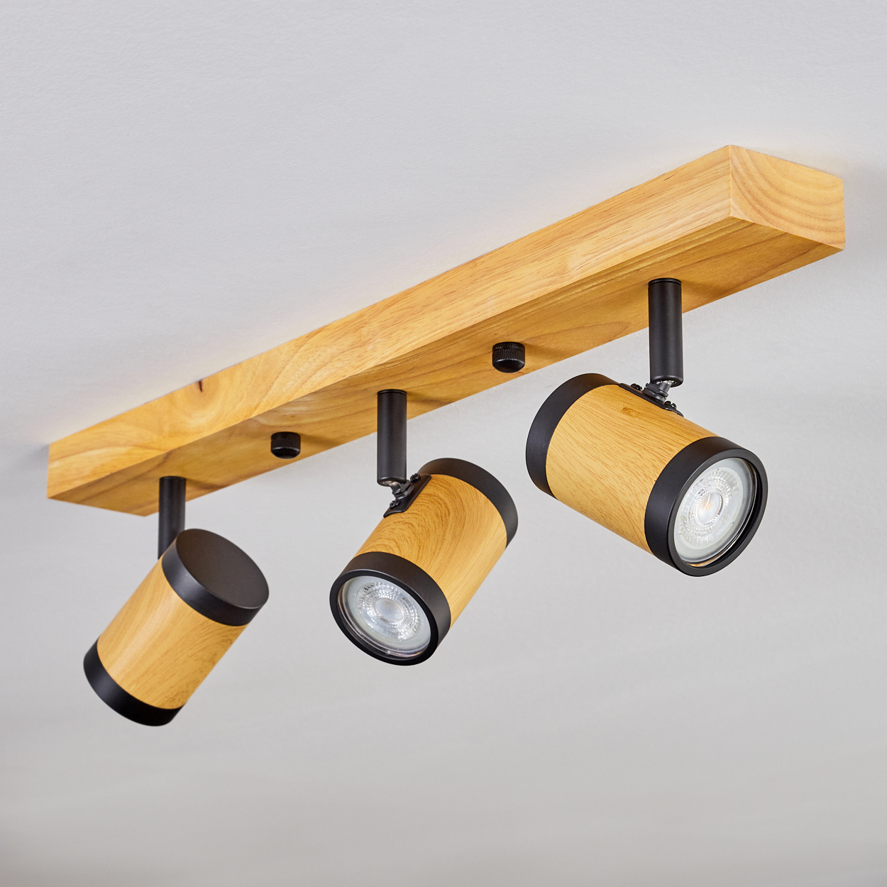 Eine Holzleuchte mit drei schwarz umrandeten Lichtern. Die Leuchte ist an einer weißen Decke montiert, die Lichter sind nach unten gerichtet.