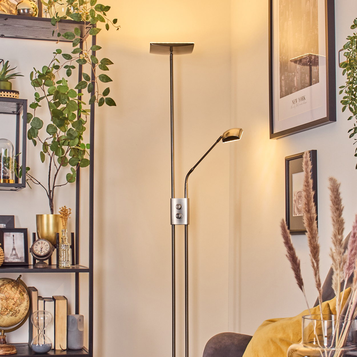 Eine moderne Stehlampe steht in einer Zimmerecke. Regale mit Deko und Pflanzen befinden sich links. Bilder hängen an der Wand.