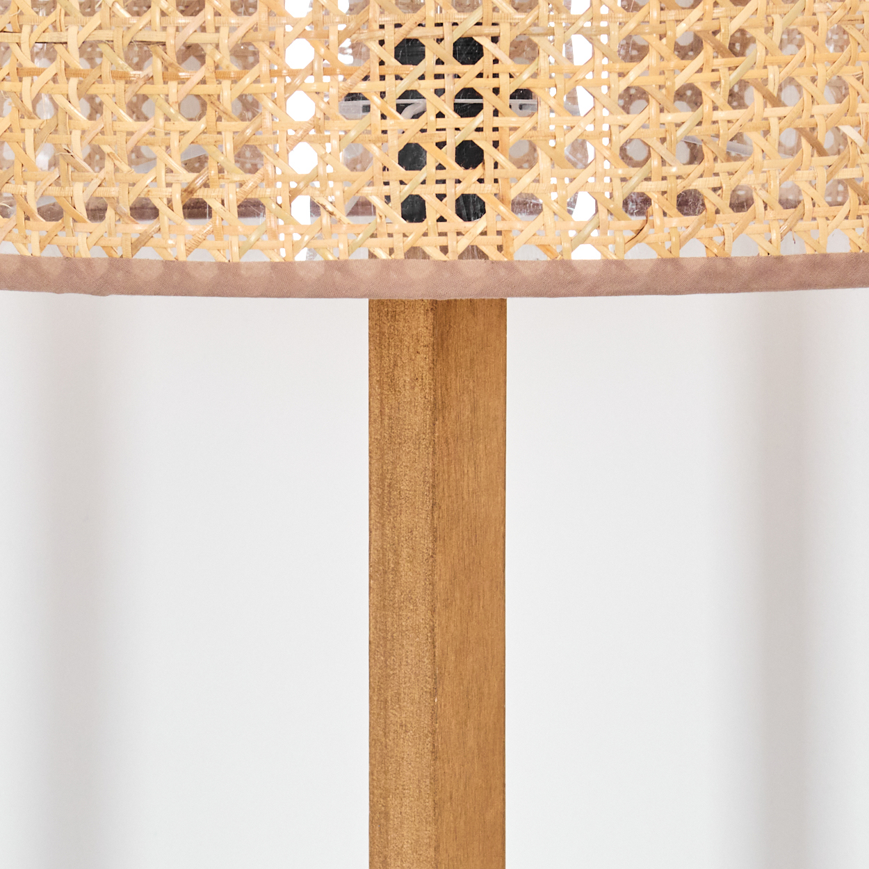 Nahaufnahme einer Lampe. Die Lampe hat einen gewebten Korbschirn mit braunem Besatz. Sie steht auf einem Holzpfosten vor weißem Hintergrund.