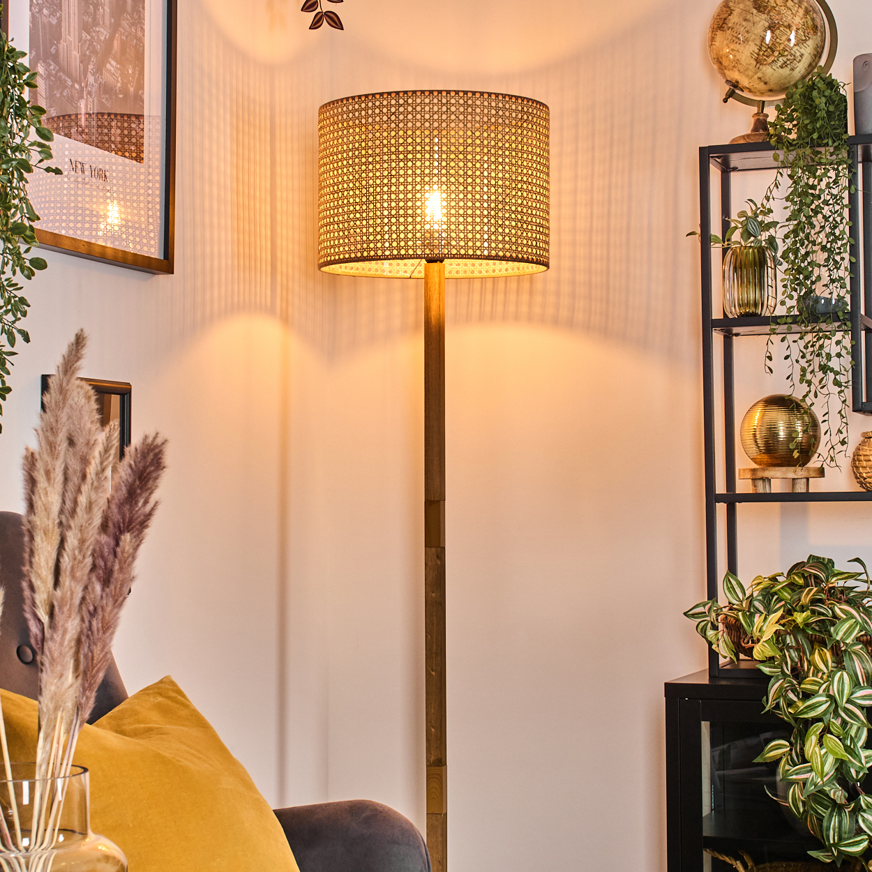 Eine beleuchtete Stehlampe mit gewebtem Schirm steht in der Ecke, neben einem Regal mit Dekoration und Pflanzen. Ein gerahmter New York-Druck hängt.