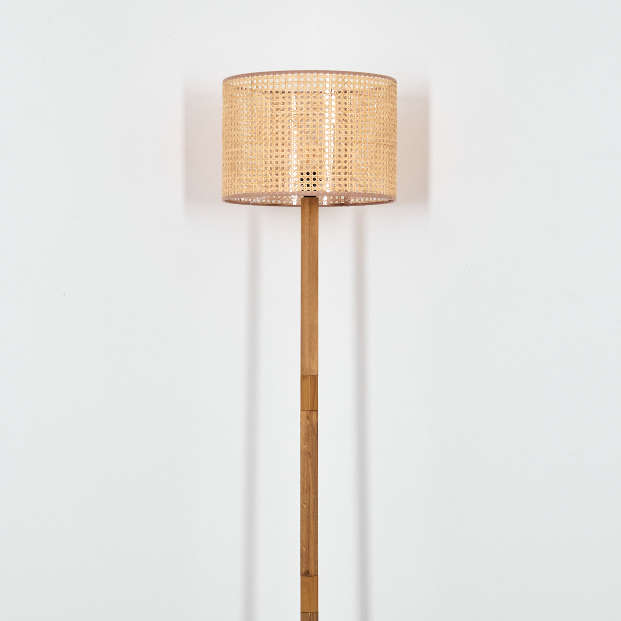 Eine hölzerne Stehlampe mit einem gewebten Lampenschirm steht vor einer weißen Wand. Der Lampenschirm ist zylindrisch und beige.