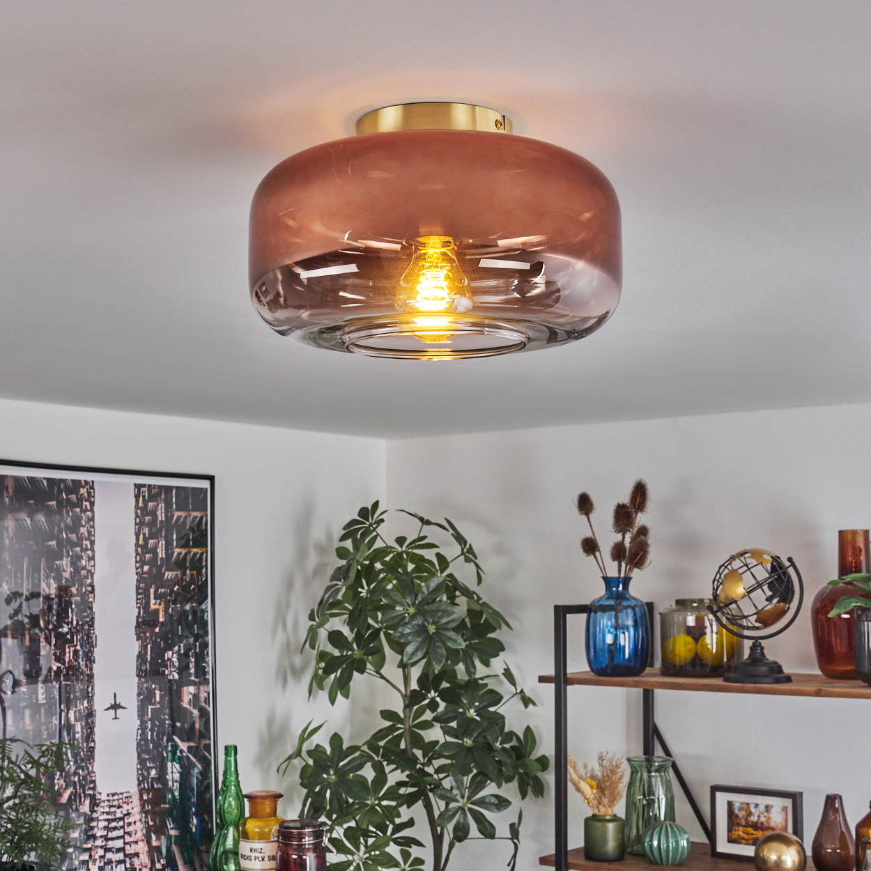 Eine Deckenlampe mit braunem Glasschirm und goldenem Sockel ist beleuchtet. Der Raum hat Pflanzen und Dekorationsartikel.