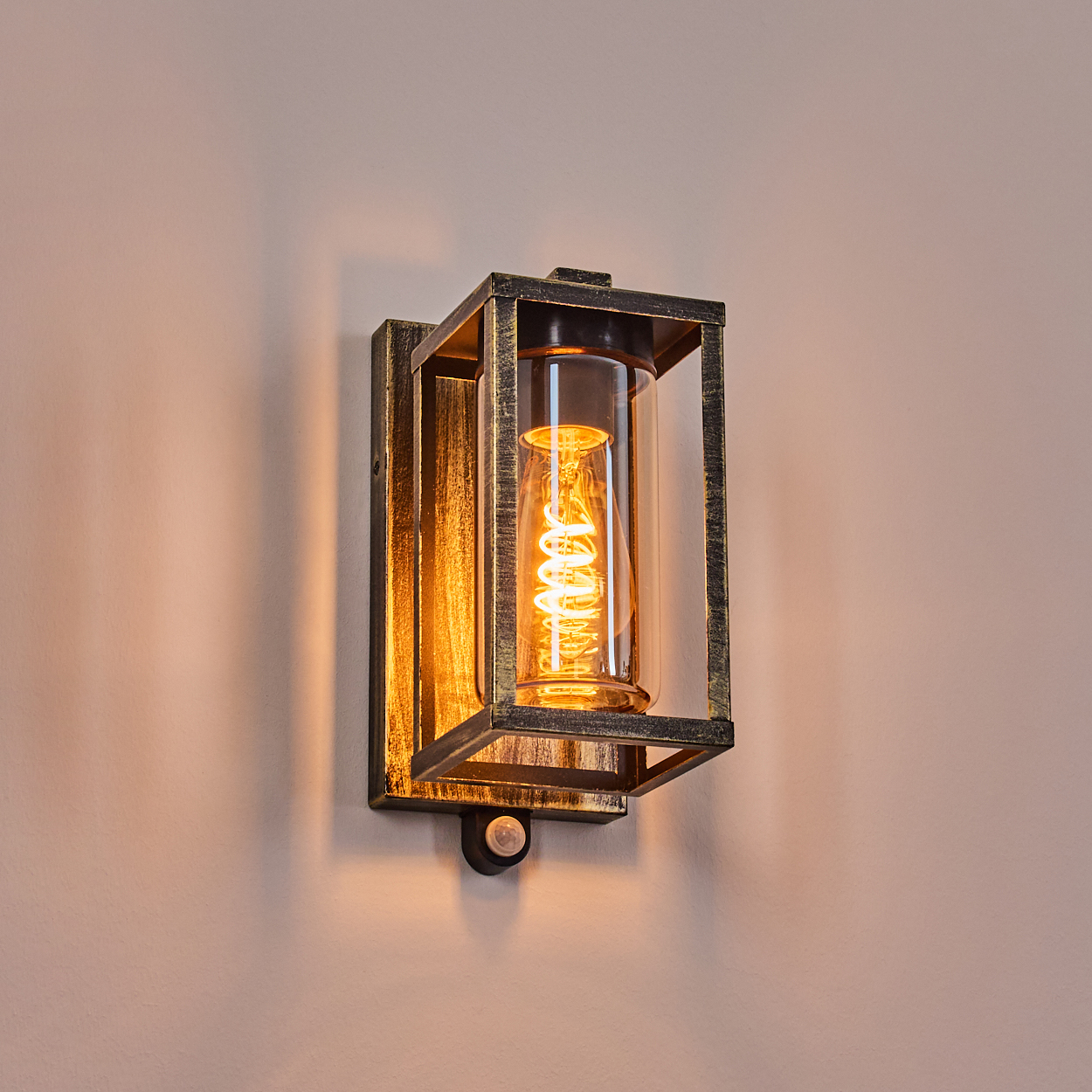 Eine Wandleuchte mit goldfarbenem Rahmen, einem Glaszylinder und einer Wendelglühbirne. Sie befindet sich an einer pfirsichfarbenen Wand.
