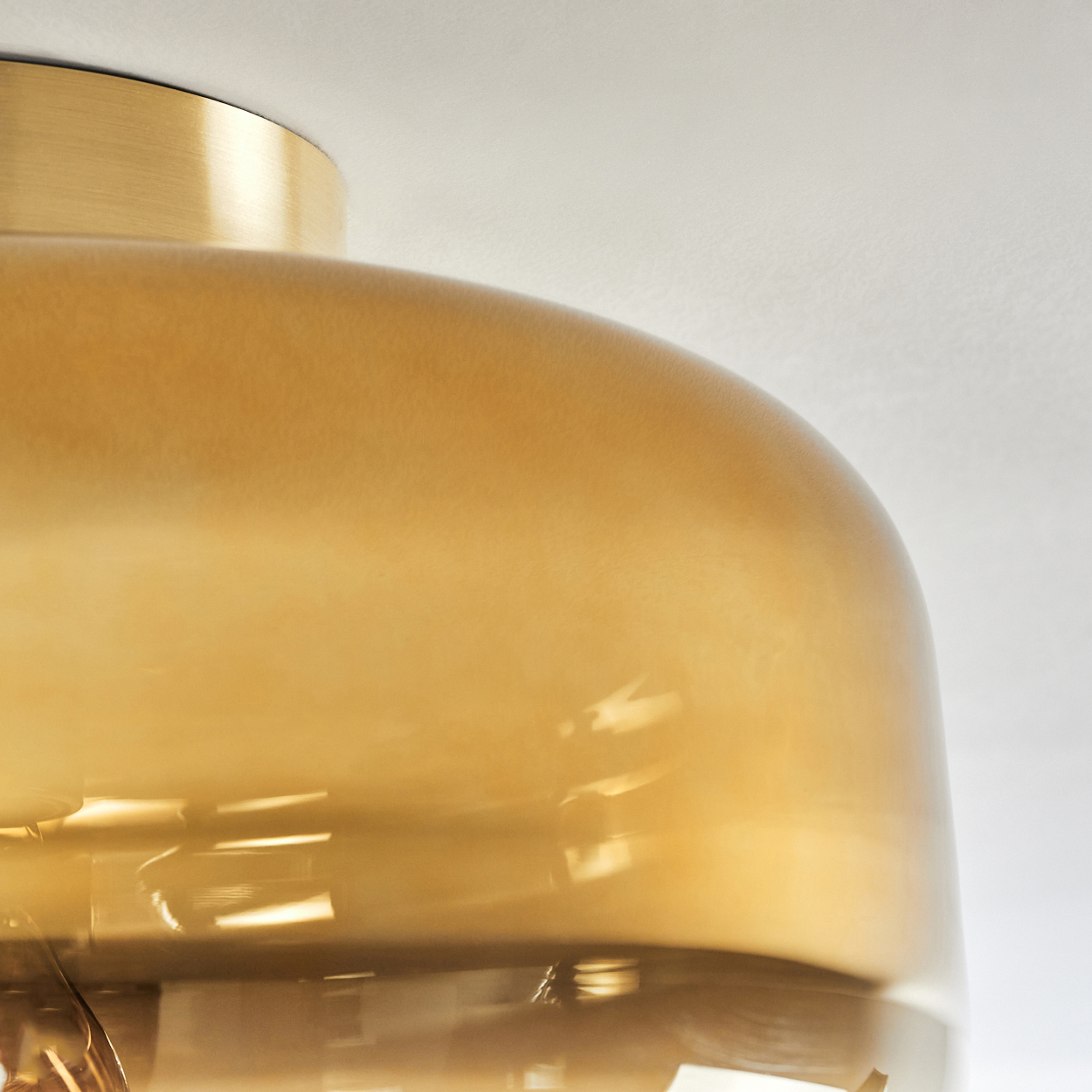 Nahaufnahme einer Leuchte. Sie hat eine goldfarbene Oberseite und einen großen, bernsteinfarbenen Glasschirm. Der Hintergrund ist eine weiße Decke.