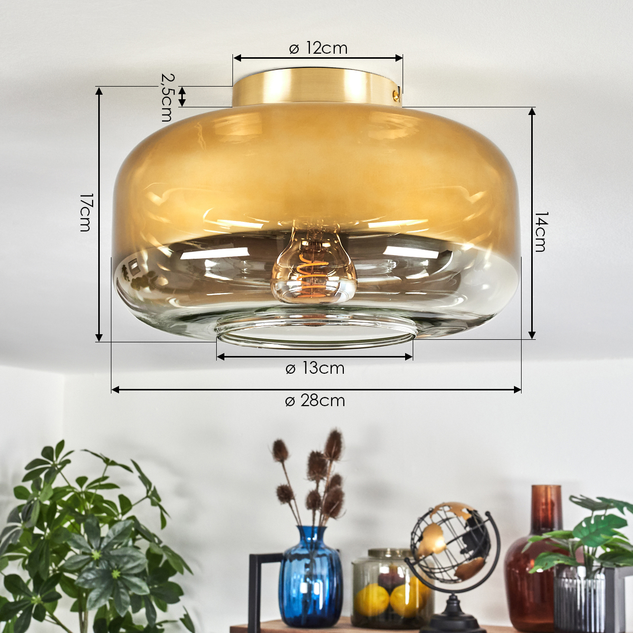 Eine goldene Deckenlampe aus Glas mit Maßangaben. Darunter ein Regal mit Pflanzen, Globus, Vasen und Zitronen im Glas.