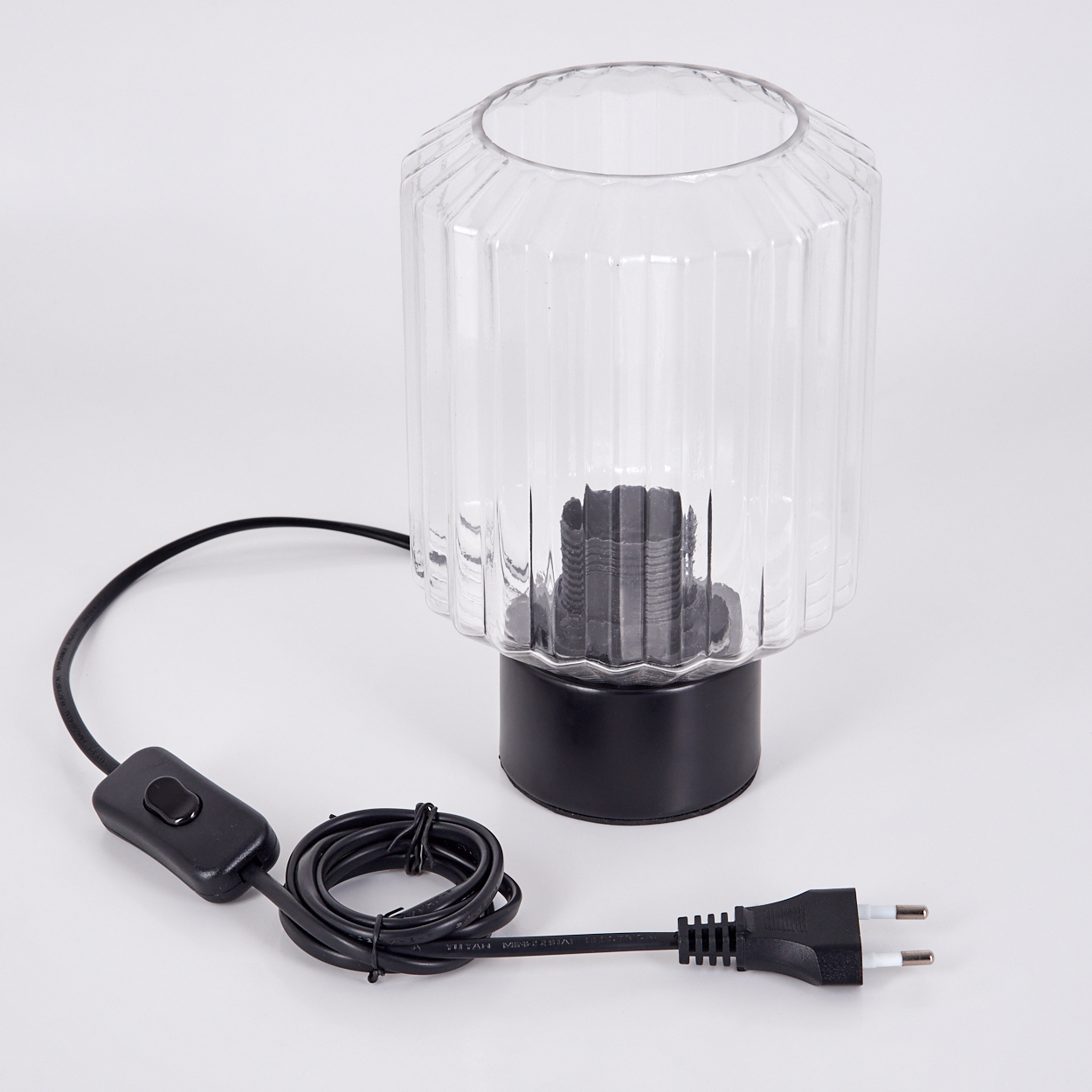 Eine klare Glaslampe mit schwarzem Sockel und Kabel. Das Kabel hat einen Schalter und Stecker. Die Lampe steht auf einer weißen Oberfläche.