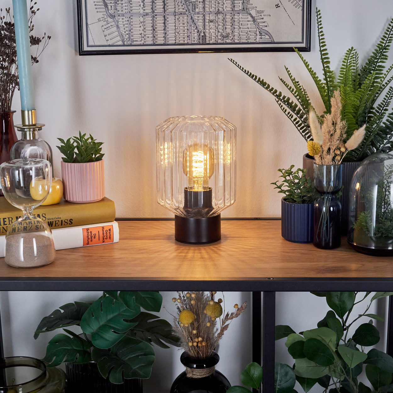 Eine beleuchtete Lampe steht auf einem Holzregal mit Pflanzen, Büchern und einer Karte. Andere Pflanzen sind unten. Hinten ist ein Kerzenhalter.