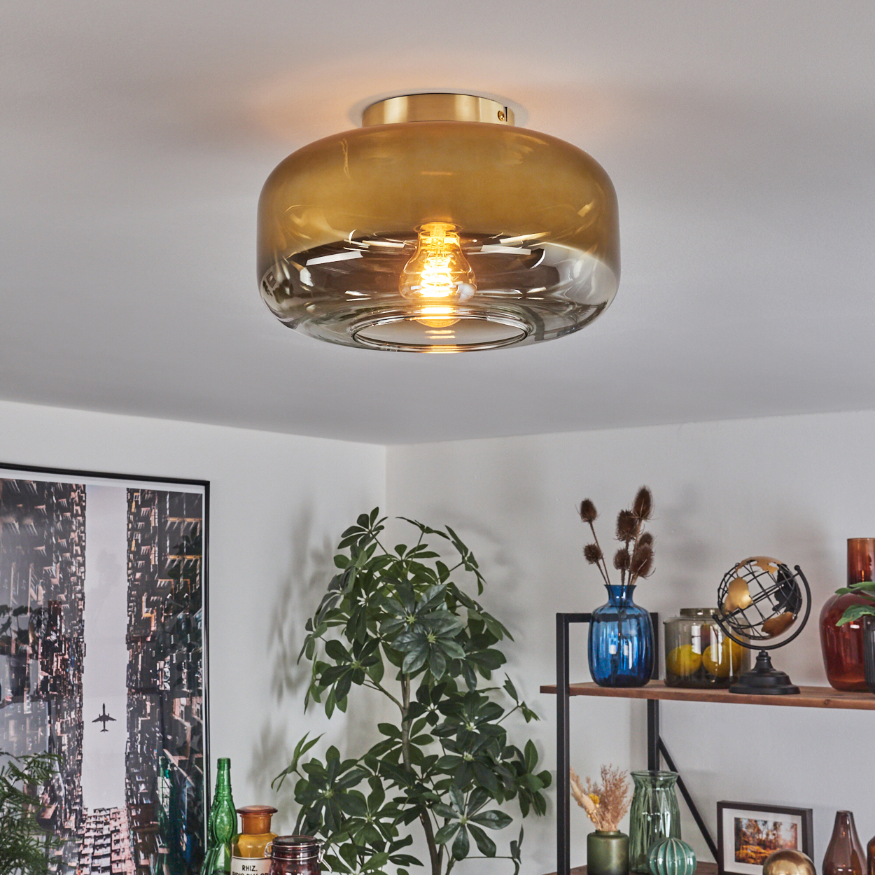 Eine Deckenleuchte mit goldfarbenem Sockel und bernsteinfarbenem Glasschirm. Eine Pflanze, Regale mit Dekoration und ein Stadtbildposter sind im Raum.