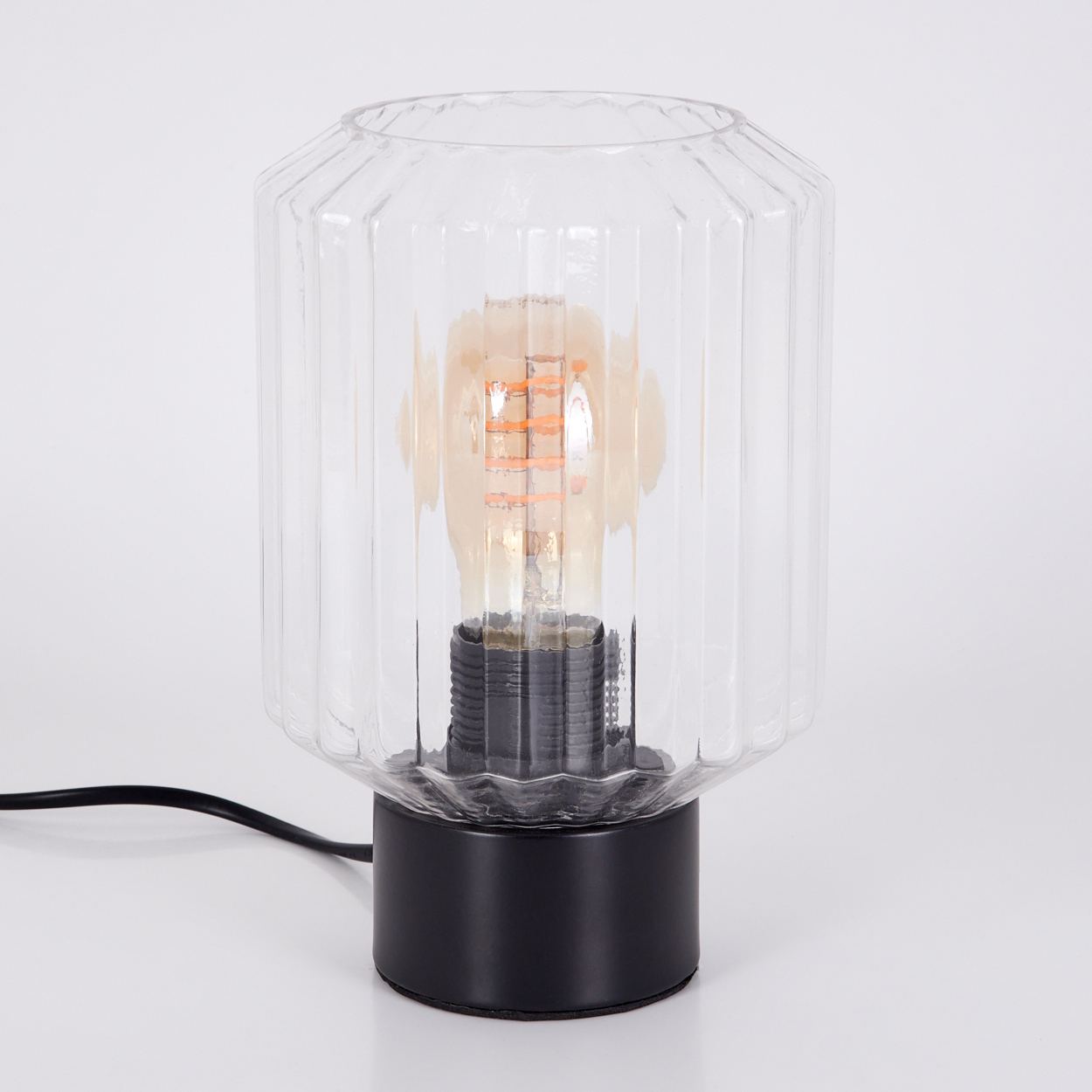 Eine Glaslampe mit schwarzem Sockel. Die Lampe hat einen transparenten, geriffelten Glasschirm und eine freiliegende Glühbirne.