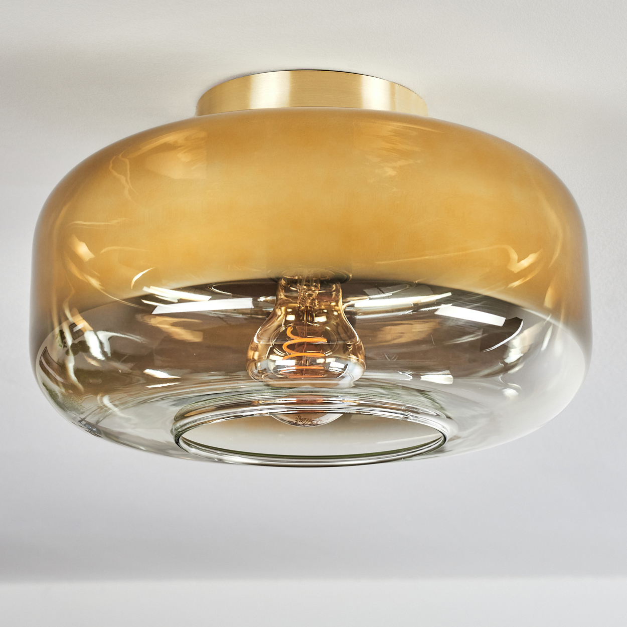 Eine moderne Deckenleuchte. Sie hat einen goldfarbenen Metallsockel und einen klaren Glasschirm mit bernsteinfarbenem Farbverlauf. Eine Glühbirne ist innen.