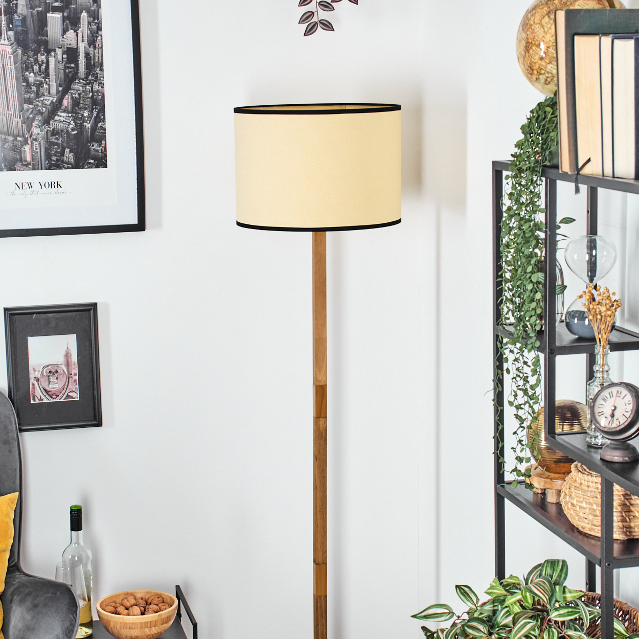 Eine Stehlampe mit beigem Schirm und Holzfuß steht neben einem schwarzen Regal mit Büchern und Pflanzen. Bilder an der Wand.