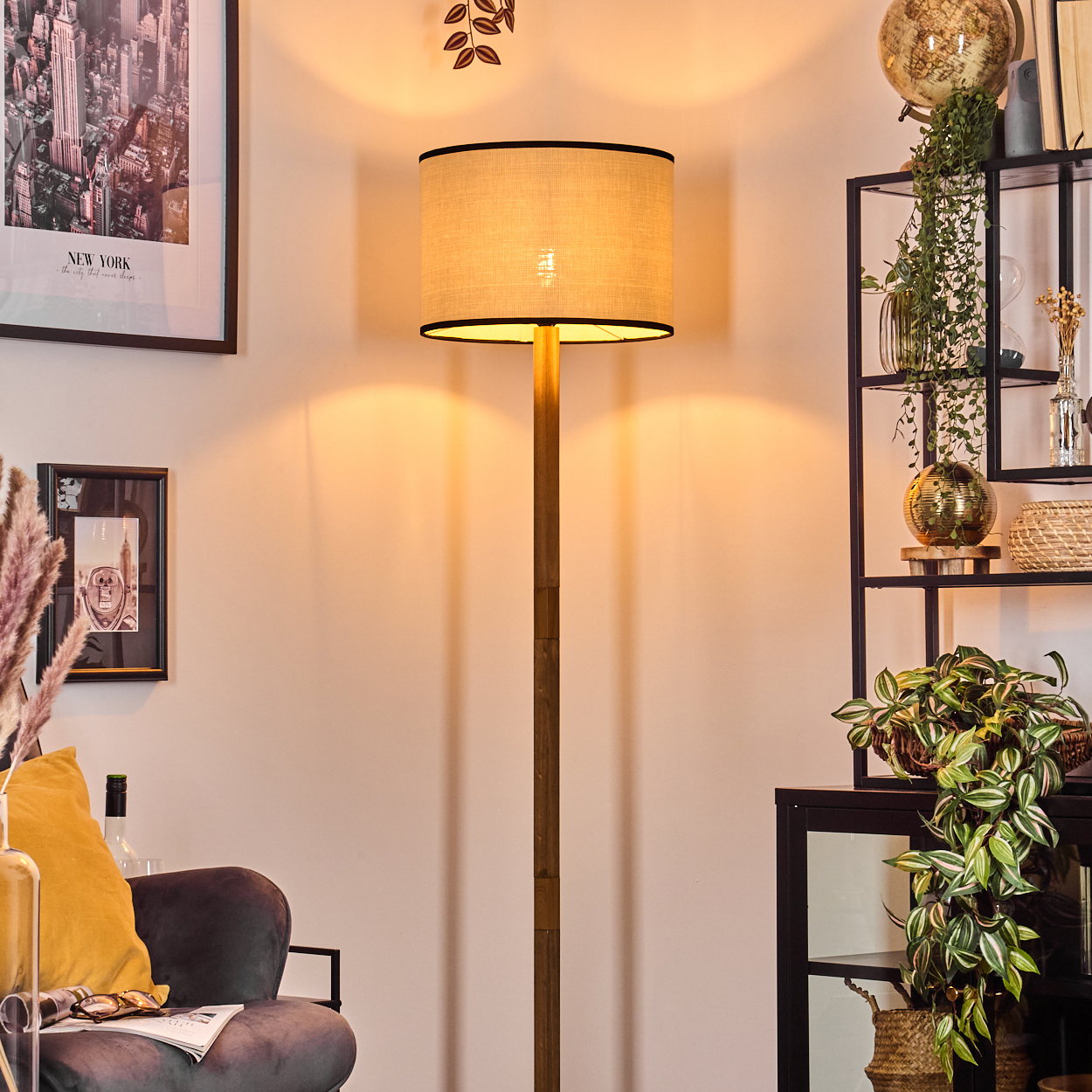 Eine beleuchtete Stehlampe steht in einem Raum. Ein New York-Bild, ein Stuhl und ein Regal mit Pflanzen und Dekor sind ebenfalls sichtbar.