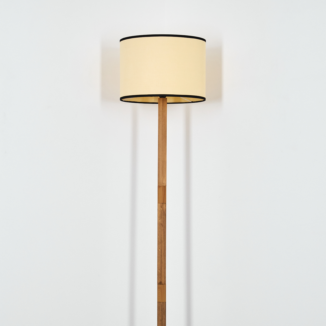 Eine Stehlampe mit beigem Schirm, schwarzem Rand und Holzstiel steht vor einer weißen Wand. Die Lampe ist zentriert.