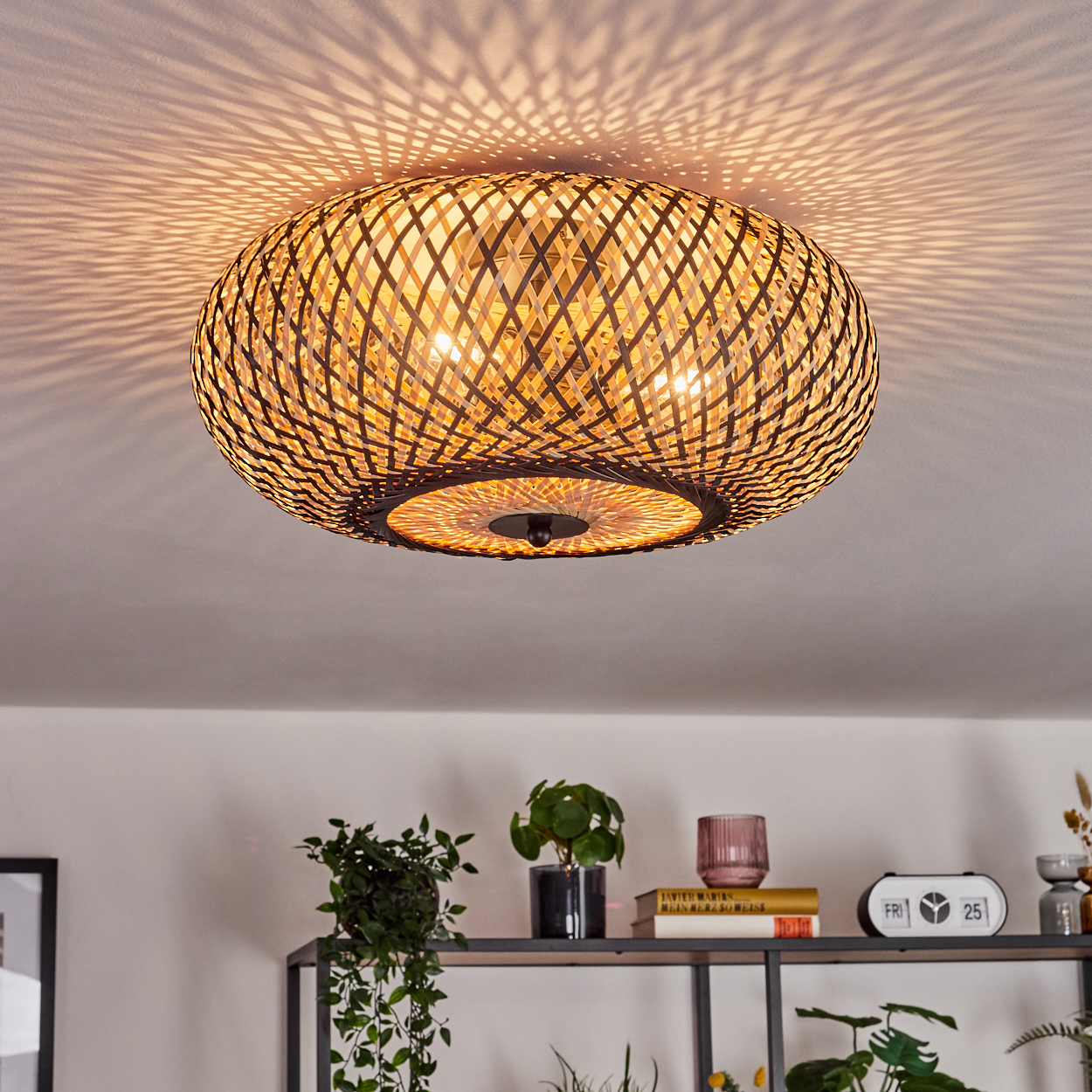Geflochtene Deckenlampe aus Rattan, die gemustertes Licht wirft. Darunter ein Regal mit Pflanzen, Büchern, einer Uhr und Dekorationsartikeln.