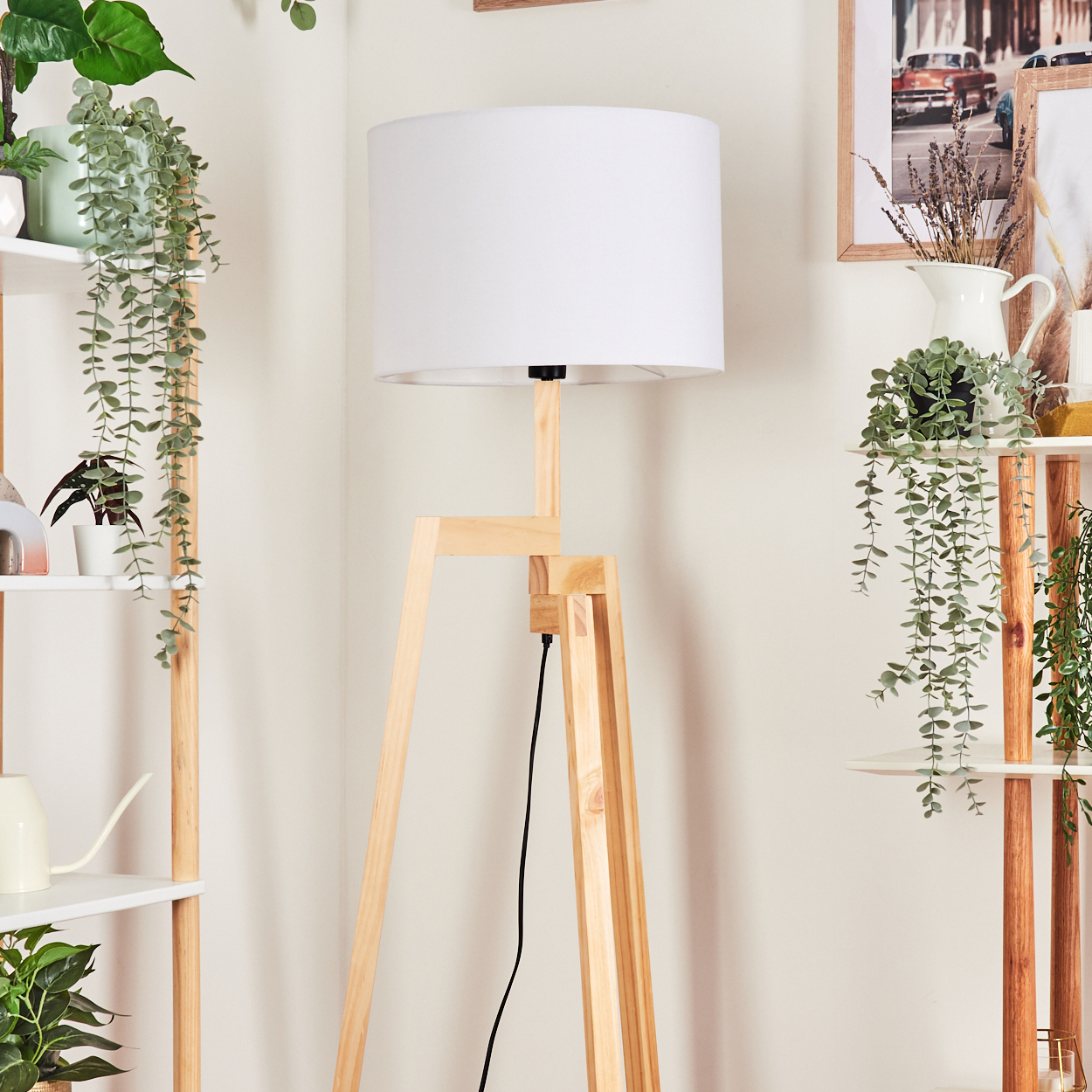 Eine hölzerne Stativ-Stehlampe mit weißem Lampenschirm steht in einem Raum mit Pflanzen auf Regalen und Wandkunst.