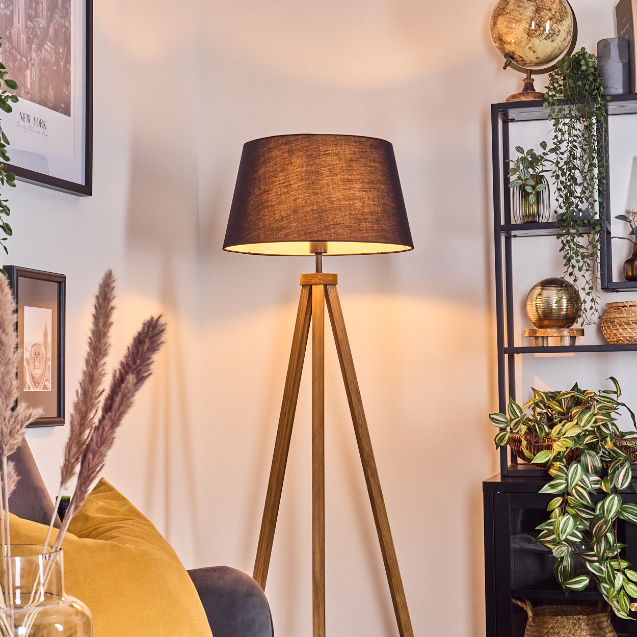 Eine hölzerne Stativlampe mit schwarzem Lampenschirm steht in einem Raum. Ein Globus, Pflanzen und ein Regal sind im Hintergrund zu sehen.
