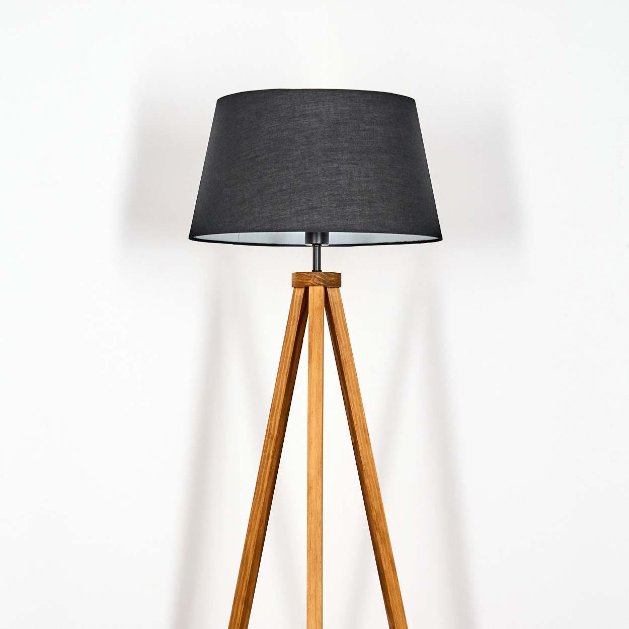 Eine hölzerne Stehleuchte mit schwarzem Lampenschirm steht vor einer weißen Wand. Die Beine der Lampe sind aus Holz.