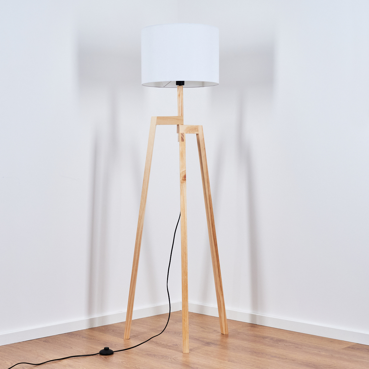 Eine Stehlampe aus Holz mit weißem Lampenschirm steht in einer Ecke. Der Boden ist aus Holz, die Wände sind weiß.