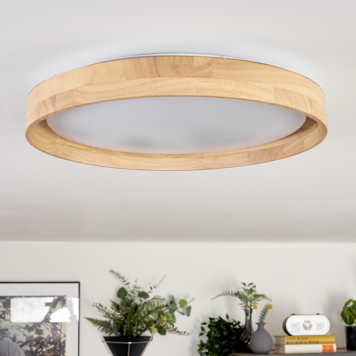 Eine kreisförmige Deckenleuchte aus Holz mit weißem Innenlicht. Darunter sind ein Regal mit Pflanzen, Büchern und einer Uhr zu sehen.