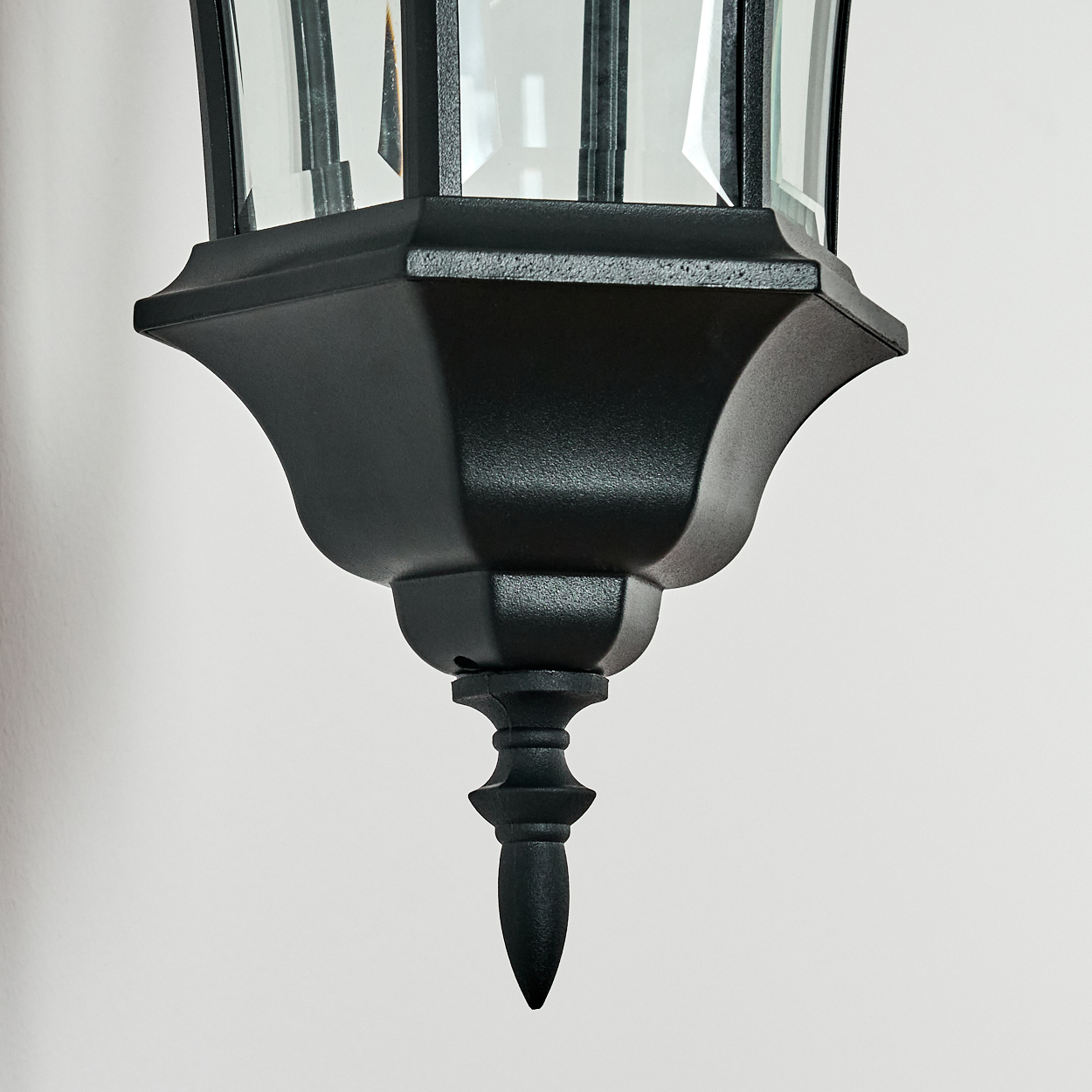 Nahaufnahme einer schwarzen Außenlaterne. Glasscheiben sind in einem schwarzen Metallrahmen sichtbar. Die Laterne hängt vor einer weißen Wand.