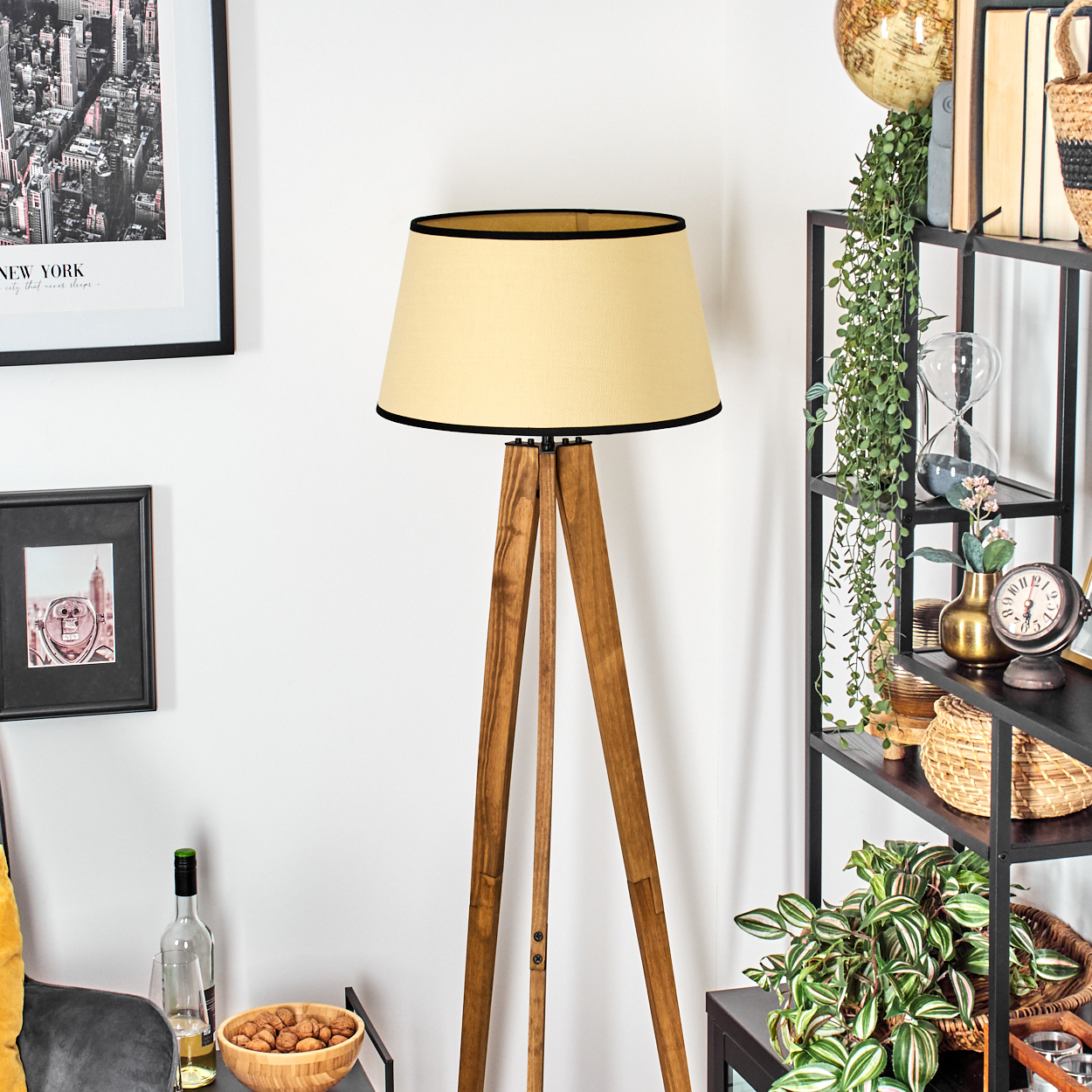 Eine Stehlampe mit beigem Lampenschirm und Holzbeinen steht vor einer weißen Wand. Regale mit Pflanzen und Dekorationen befinden sich rechts.