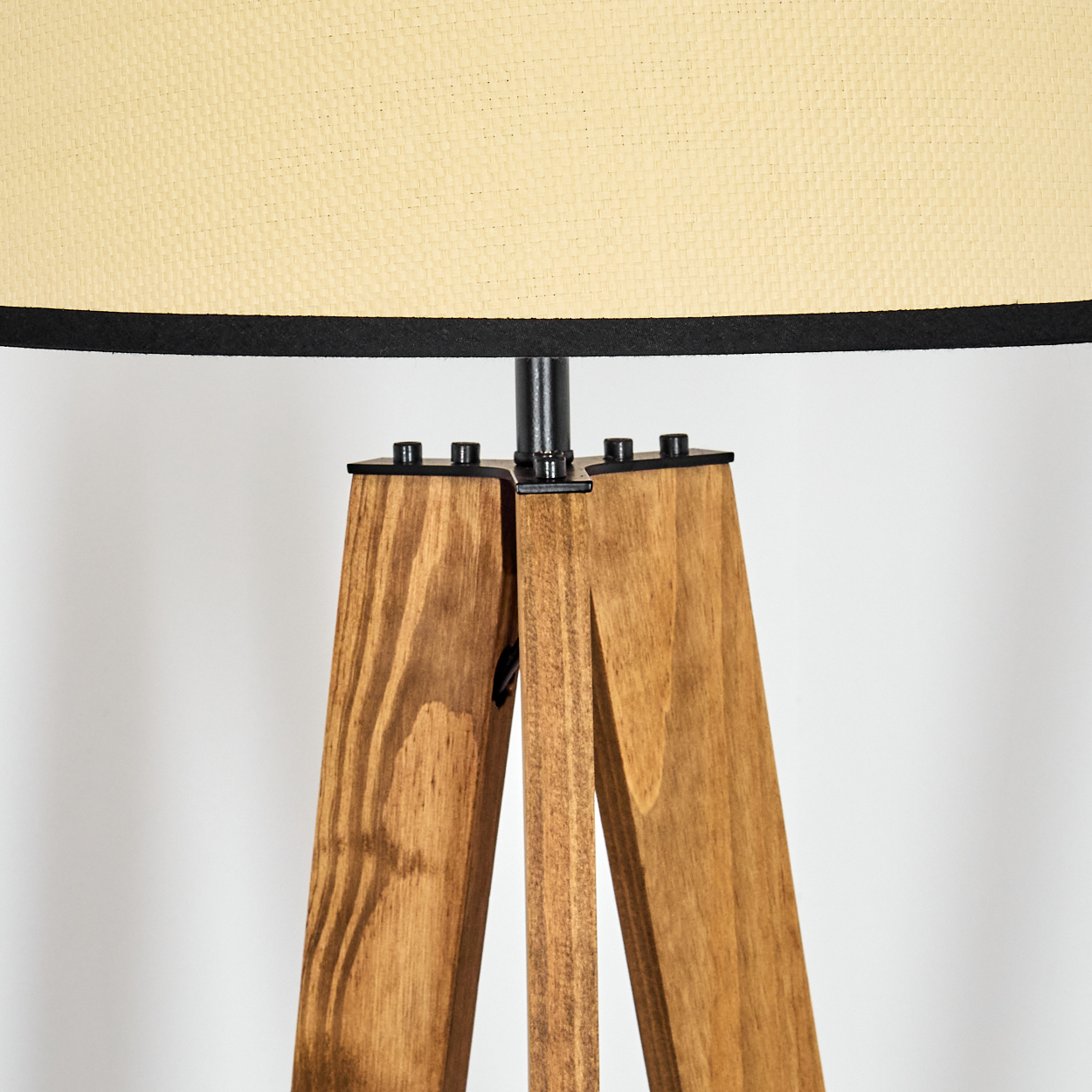 Eine Stehlampe mit Holzstativ, beigem Schirm und schwarzem Rand. Die Beine sind aus Holz, der Hintergrund ist weiß.