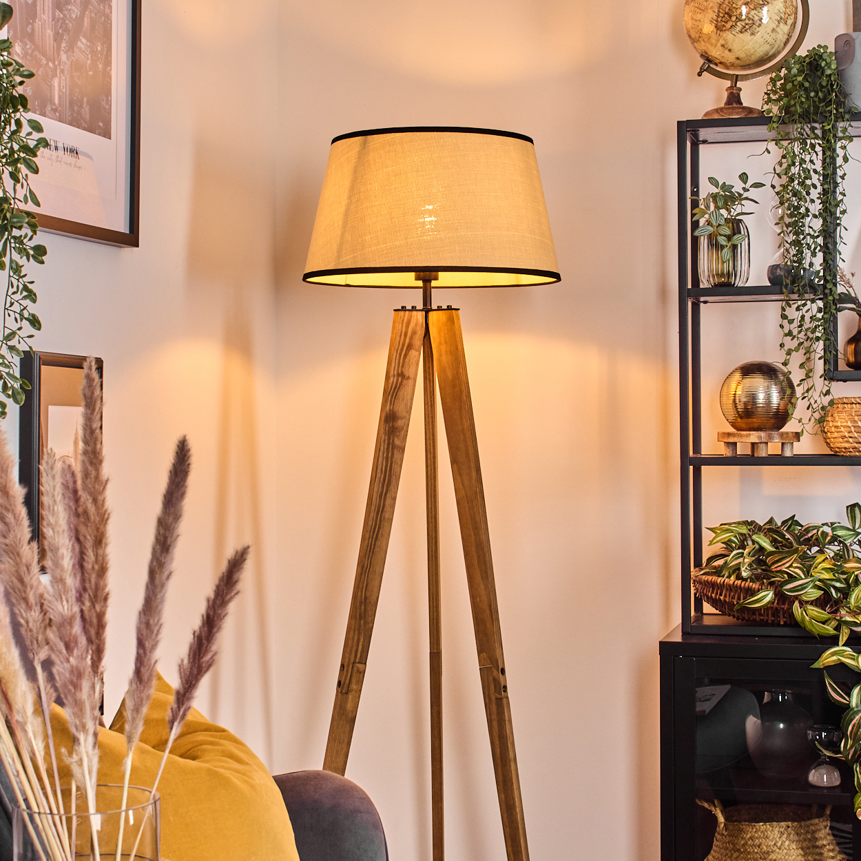 Eine hölzerne Stativ-Stehlampe mit beigem Schirm steht neben einem schwarzen Regal mit Pflanzen und einem Globus. Es gibt ein gelbes Kissen.