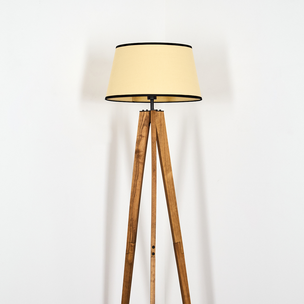 Eine hölzerne Stativ-Stehlampe mit beigem Lampenschirm und schwarzer Einfassung steht vor einer weißen Wand.