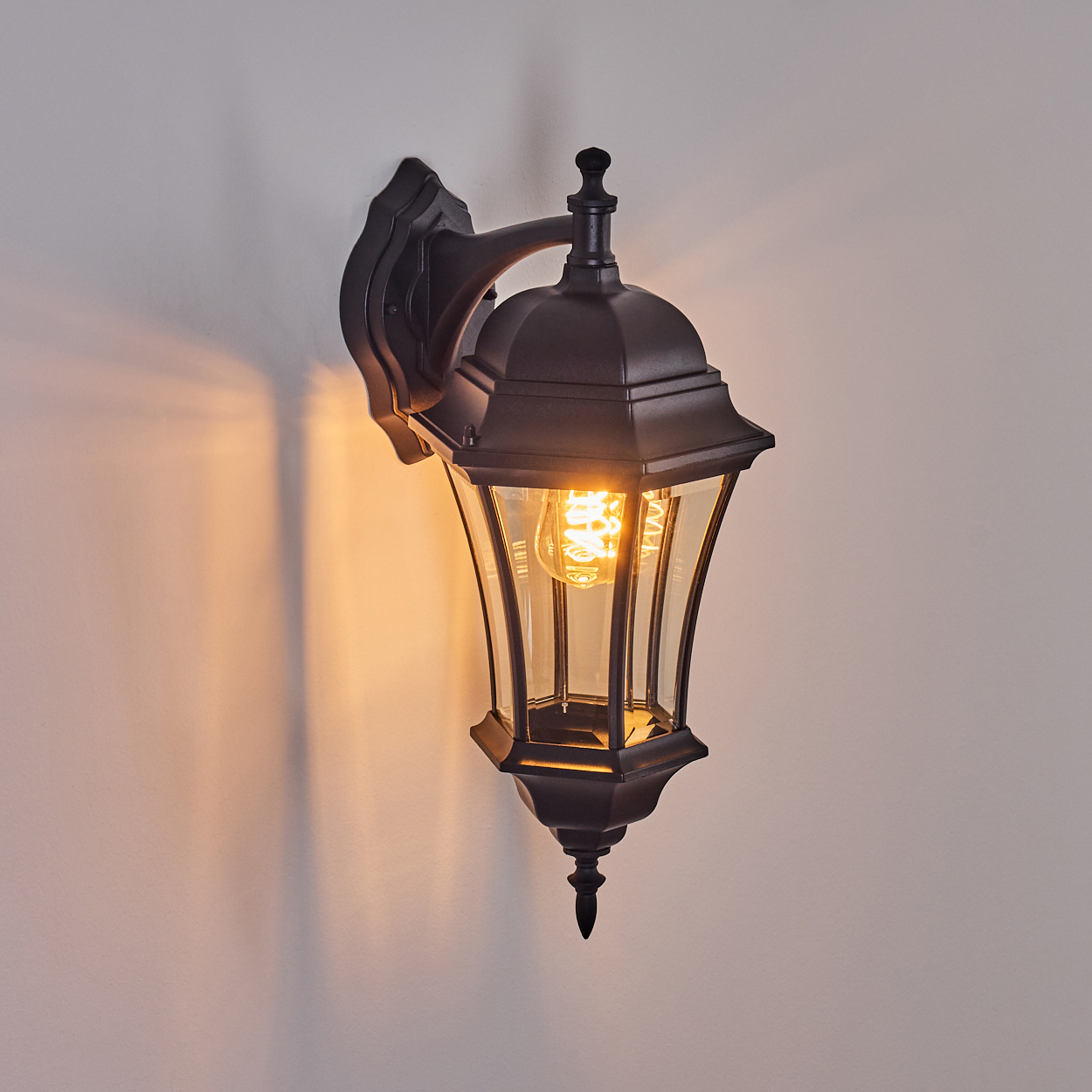 Eine schwarze Außenwandleuchte mit einer leuchtenden Glühbirne ist an einer hellgrauen Wand befestigt. Die Lampe hat ein klassisches Laternen-Design.