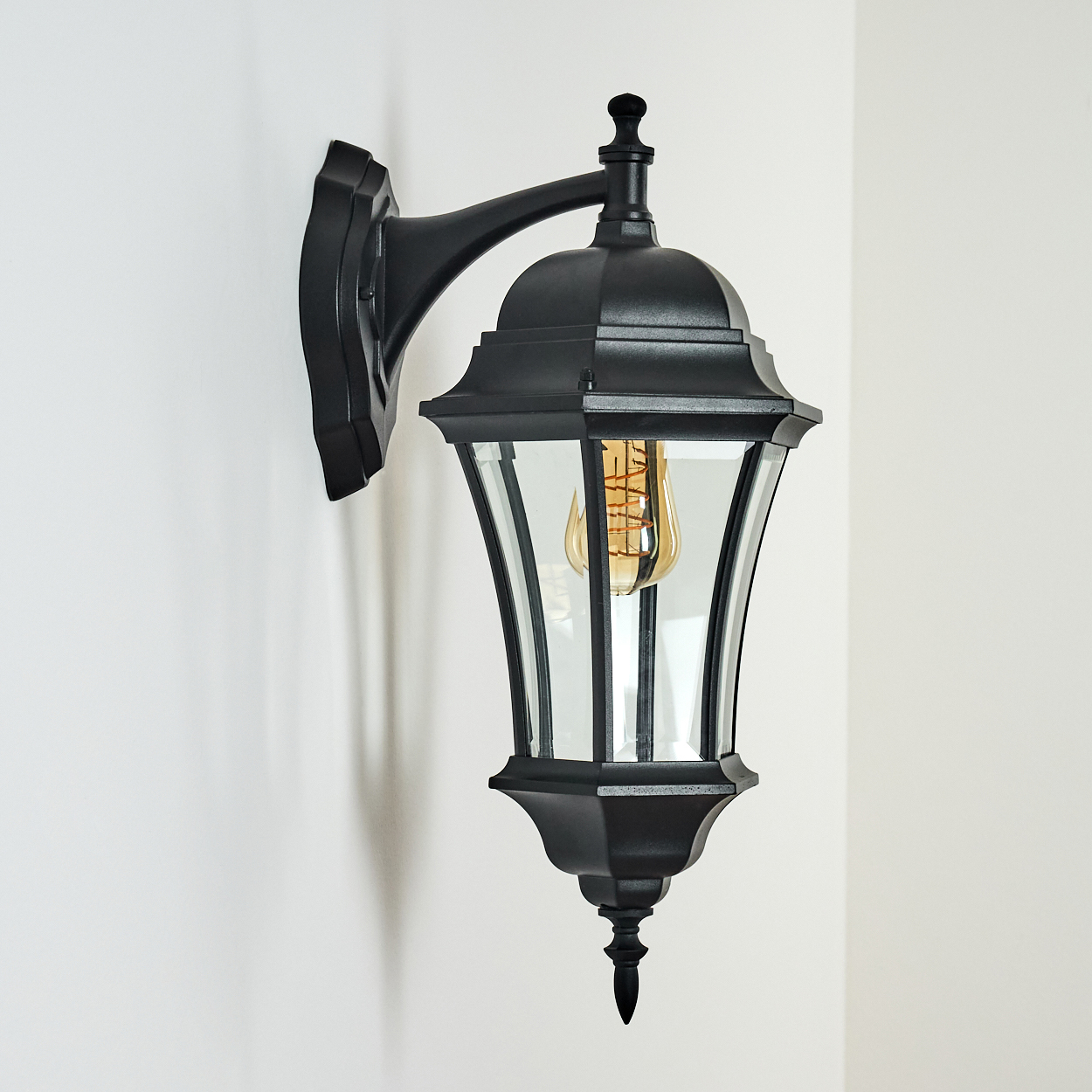 Schwarze Außenwandleuchte mit Glasscheiben und sichtbarer Glühbirne, an einer weißen Wand montiert. Die Lampe hat ein elegantes Design.